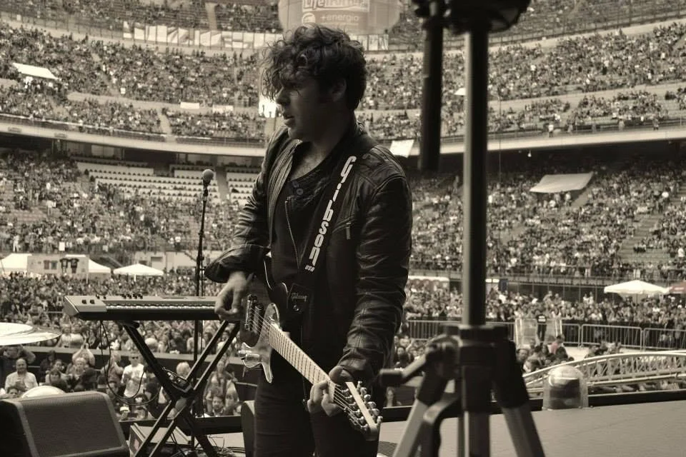 A male musician on stage at a concert with a large crowd in a stadium, holding an electric guitar and standing near a keyboard and microphone stand.