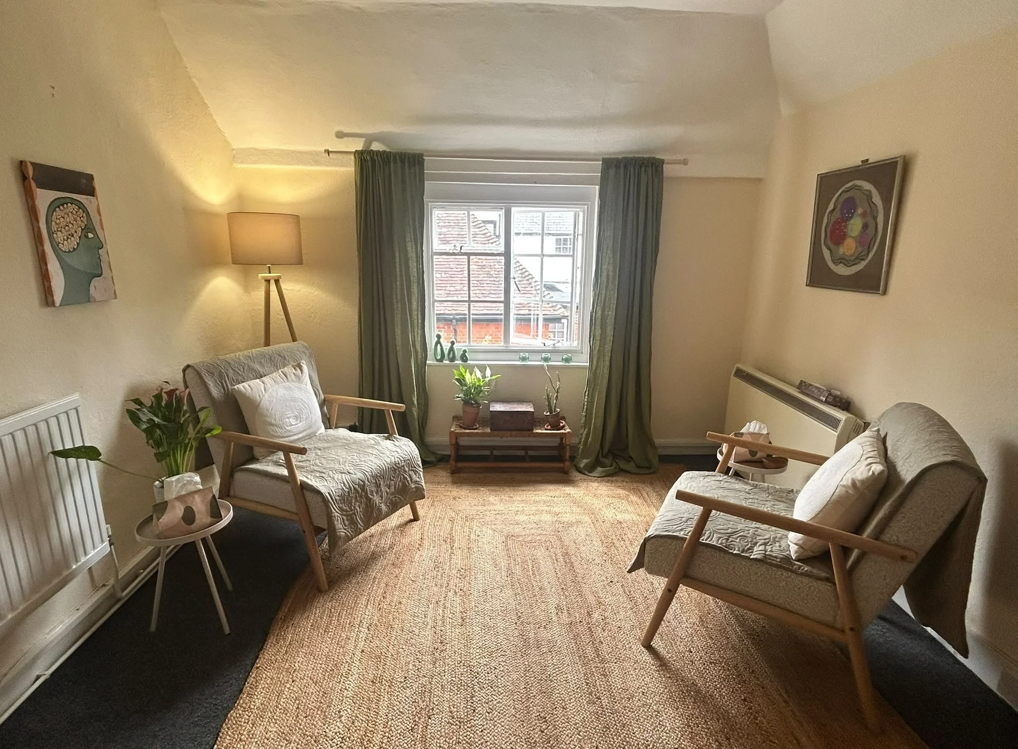 Warm and peaceful therapy room at Rooftops in Battle, East Sussex, featuring two comfortable armchairs, a natural jute rug, warm lighting, house plants, olive green curtains, and artwork on the walls.