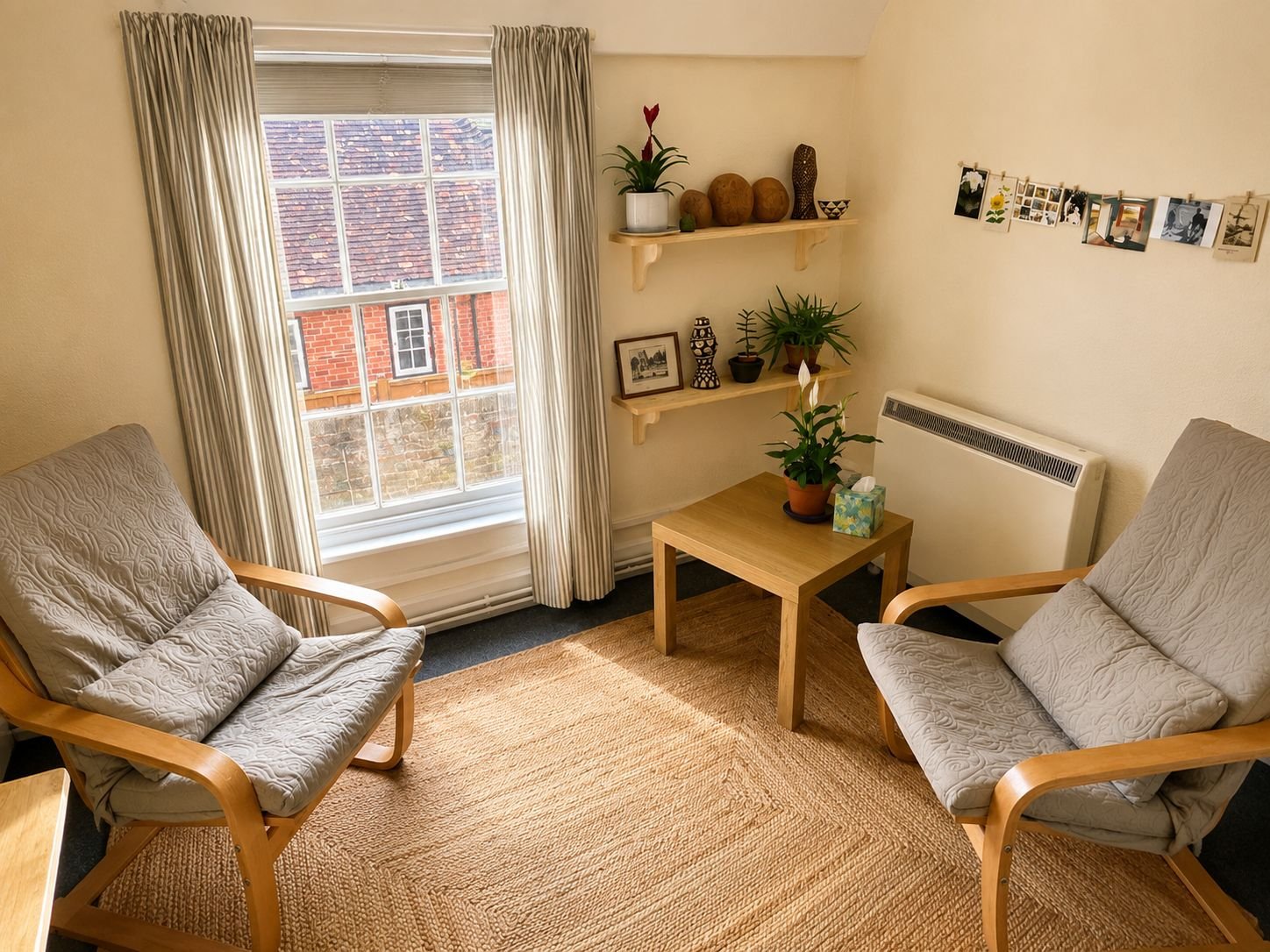 A cozy living room corner with two beige armchairs with patterned fabric and wooden frames, a small wooden table with a potted plant and a box of tissues, a window with beige curtains, and two wall-mounted shelves with decorative items and framed photos.