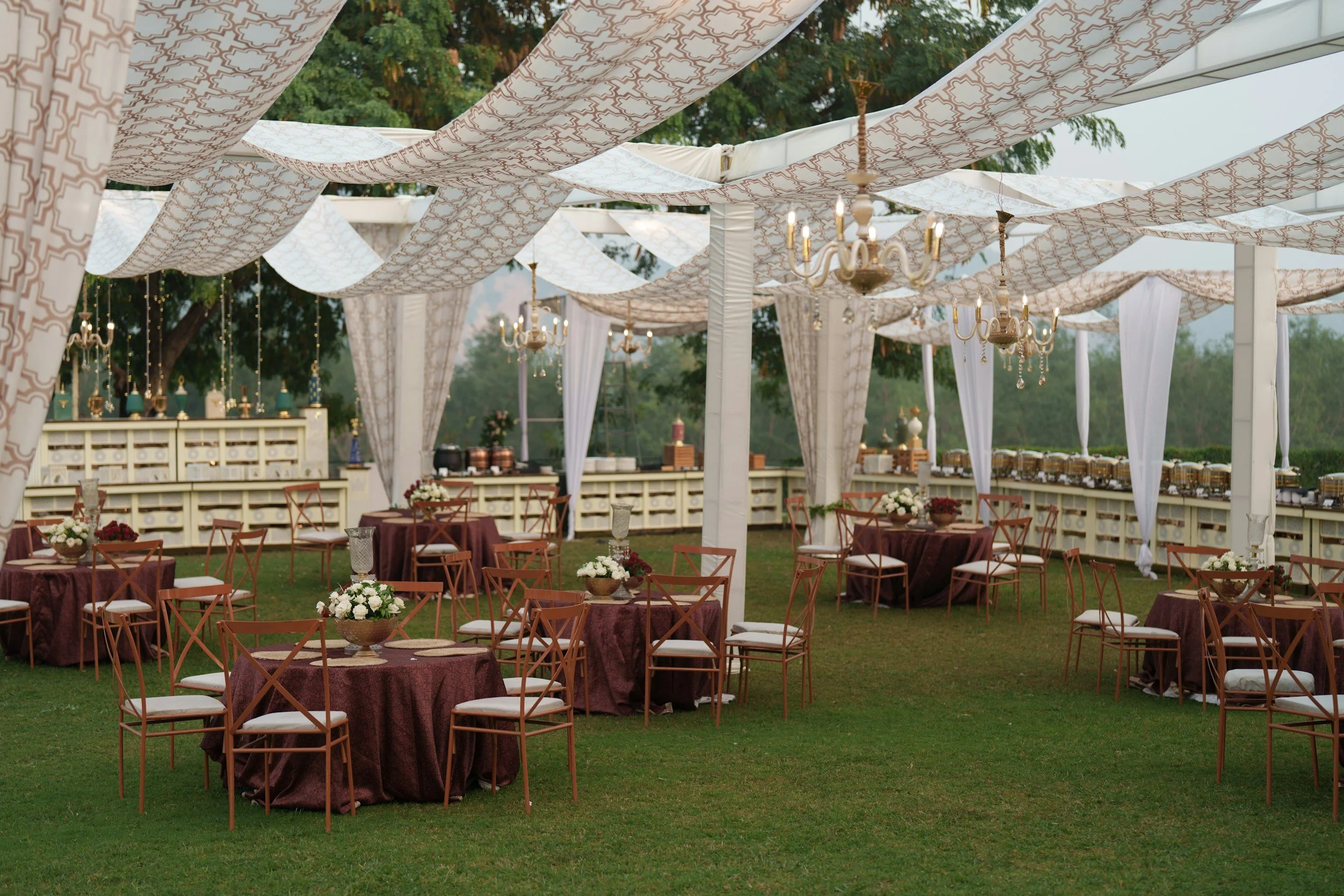 Outdoor event setup with round tables covered in maroon tablecloths, decorated with white floral centerpieces, surrounded by wooden chairs with white cushions. Overhead, white patterned fabric drapes are suspended and crystal chandeliers hang from a 