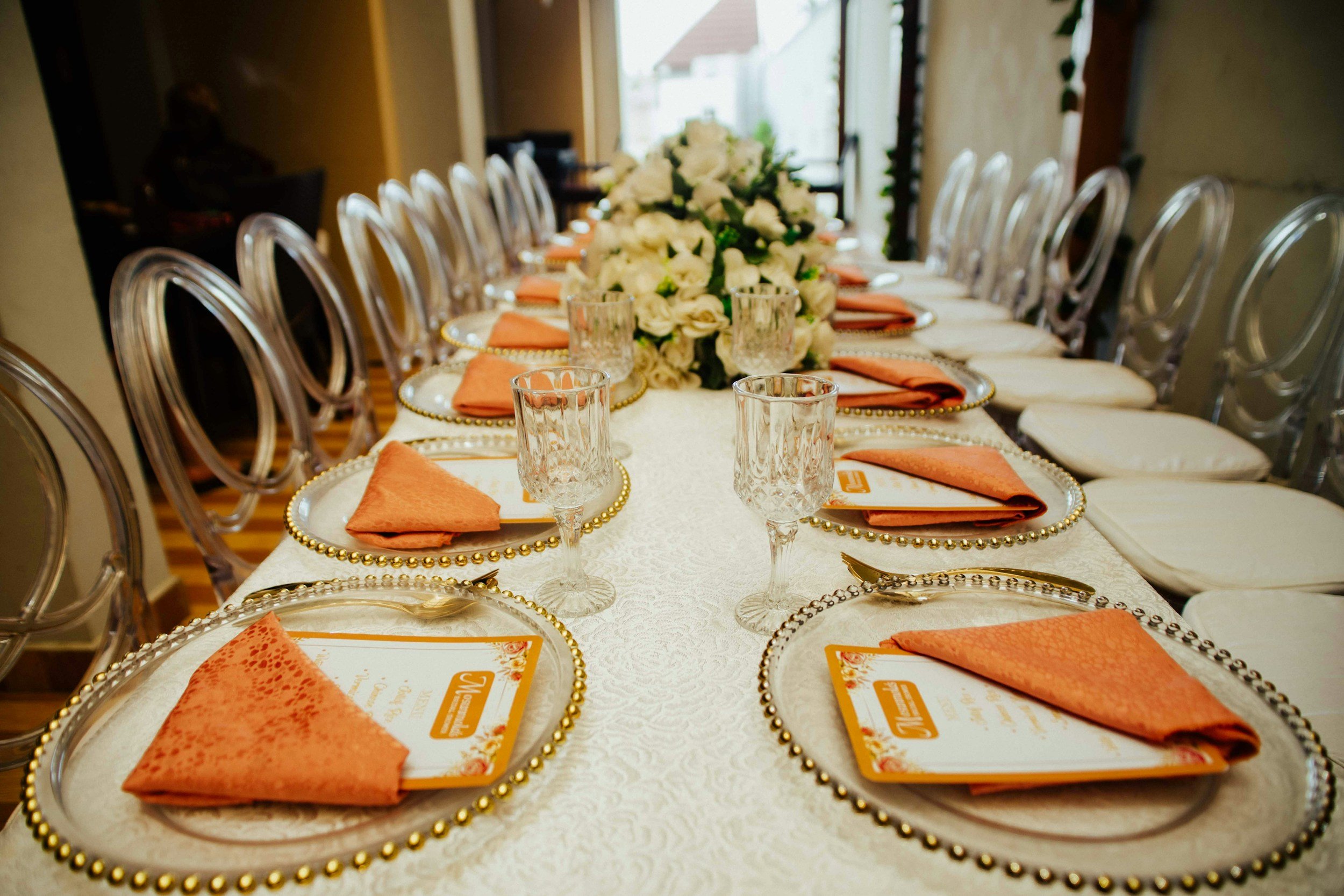 A long elegantly set dining table with white tablecloth, gold bead-edged chargers, orange cloth napkins, clear vintage glasses, and a large floral centerpiece of white roses and greenery in a bright, well-lit room.