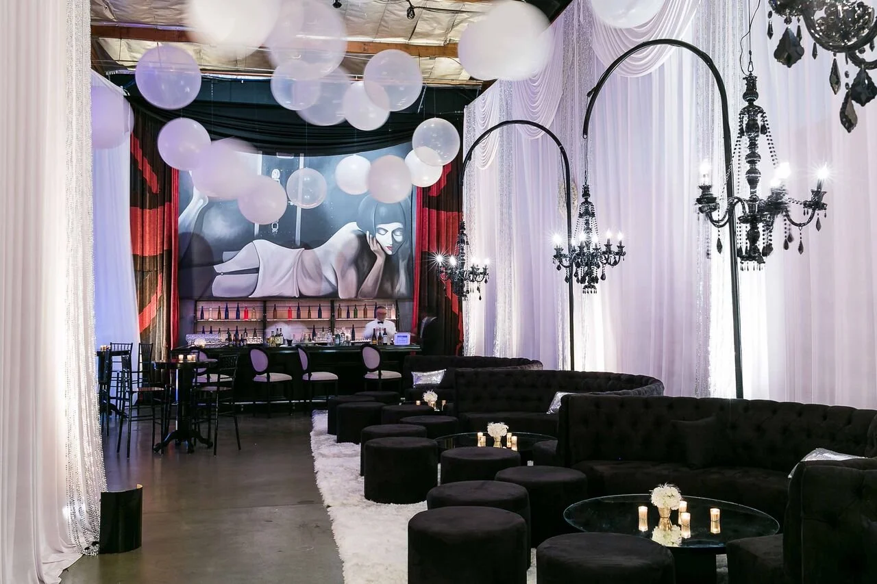 Elegant lounge with black velvet seating, chandeliers, and white curtains, featuring a large mural of a reclining woman and a bar with colorful bottles in the background.