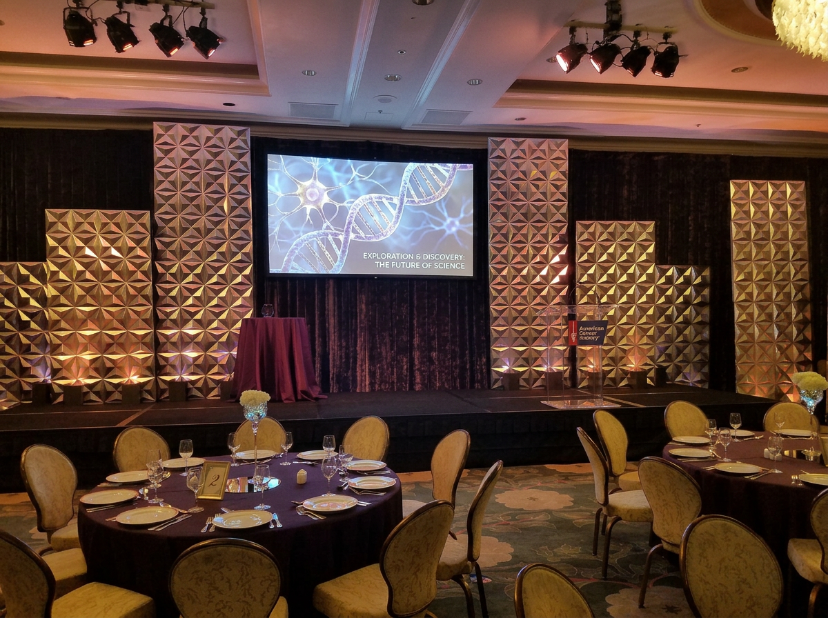 Event banquet room set for dinner with multiple round tables, each with place settings, glasses, and floral centerpieces. The stage features a large screen displaying a DNA strand and the words "Exploration & Discovery: The Future of Science," with g