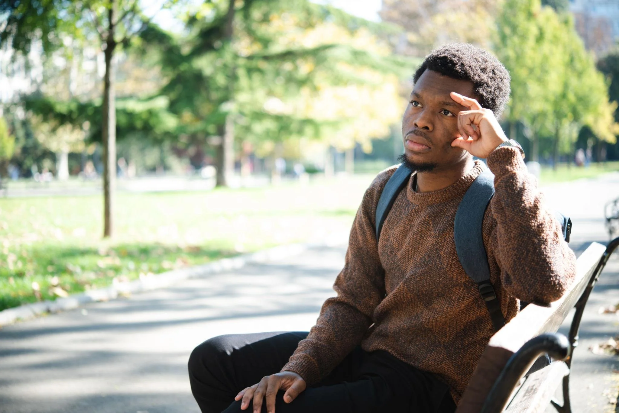 A man sitting on a park bench, looking thoughtful with his hand on his forehead, surrounded by trees and greenery.