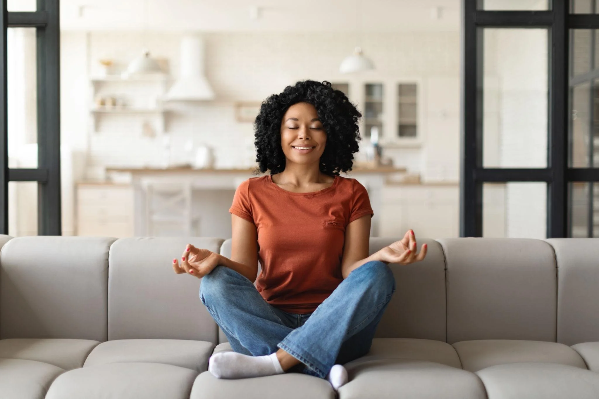 A woman with curly black hair sitting cross-legged on a gray couch in a modern kitchen, meditating with her eyes closed and a slight smile.