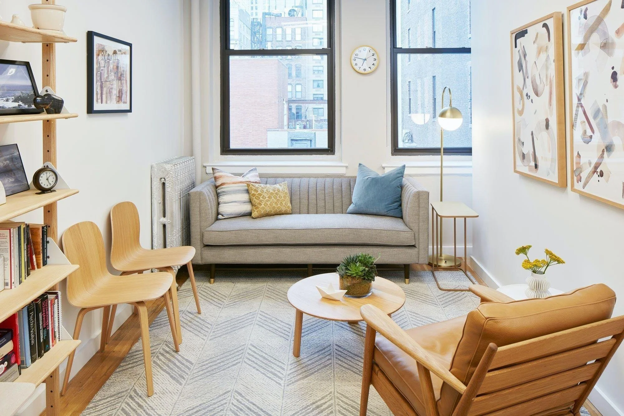 Living room with a gray sofa, wooden chairs, artwork on white walls, a round coffee table with a plant, a bookshelf with books, large windows, and a gold floor lamp.