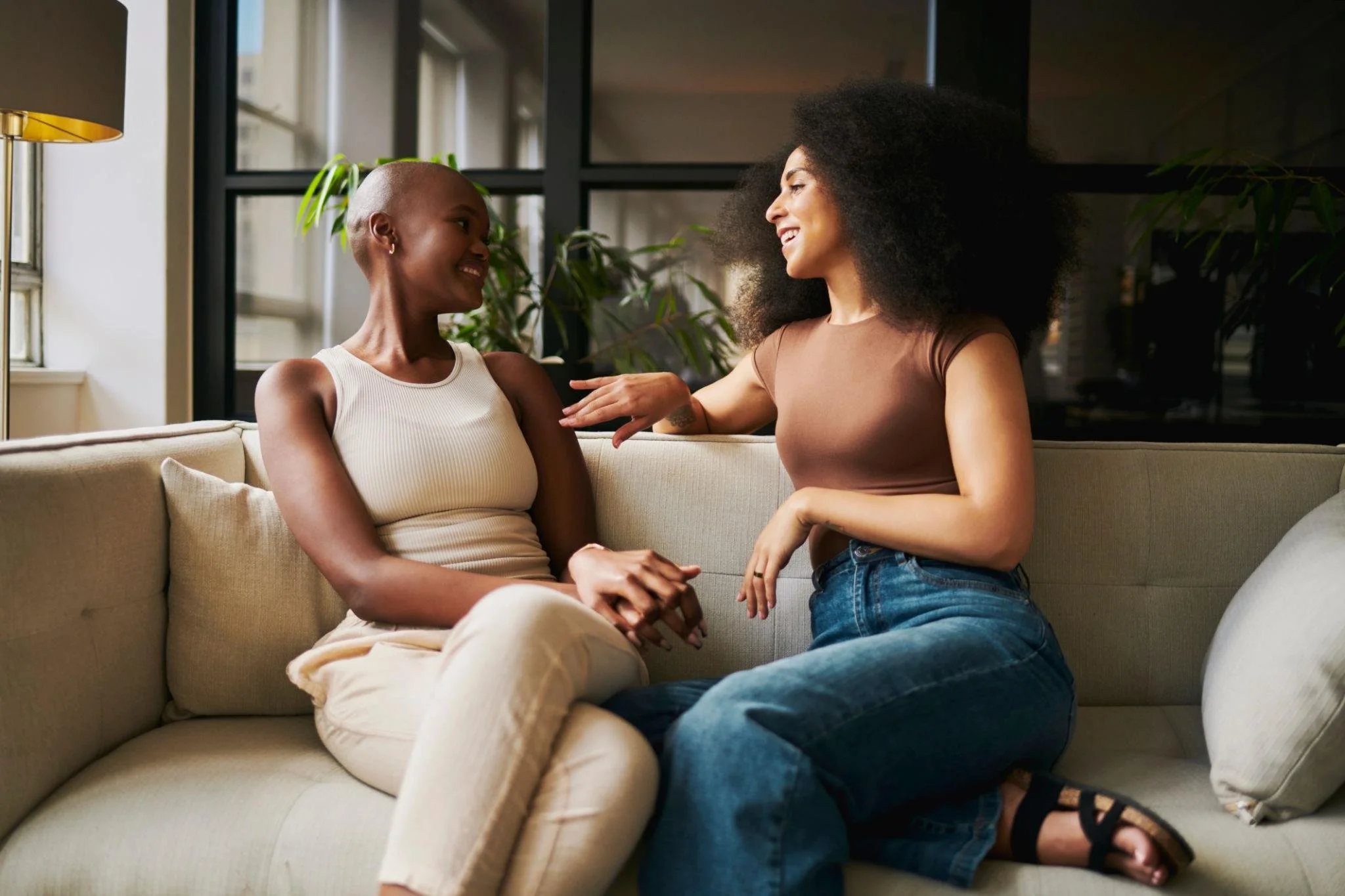 Two women sitting on a beige couch in a well-lit room, smiling and engaging in conversation, with plants and large windows in the background.