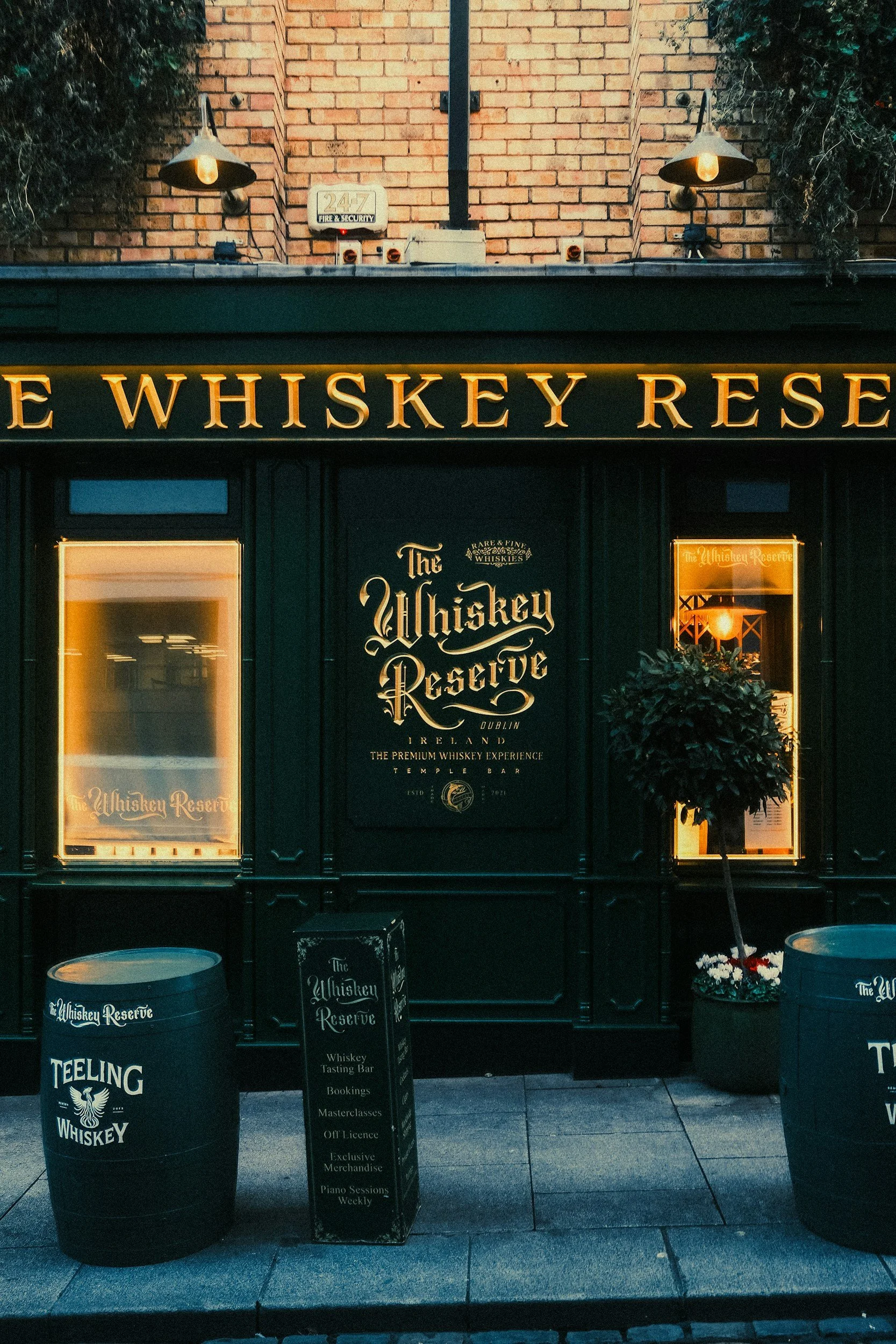 Storefront for The Whiskey Reserve bar, with illuminated sign, display windows, black exterior, and standing chalkboard signs outside listing services like tasting bar and bookings. There are two barrels outside near a small potted plant, and warm lighting inside the bar can be seen through the windows.