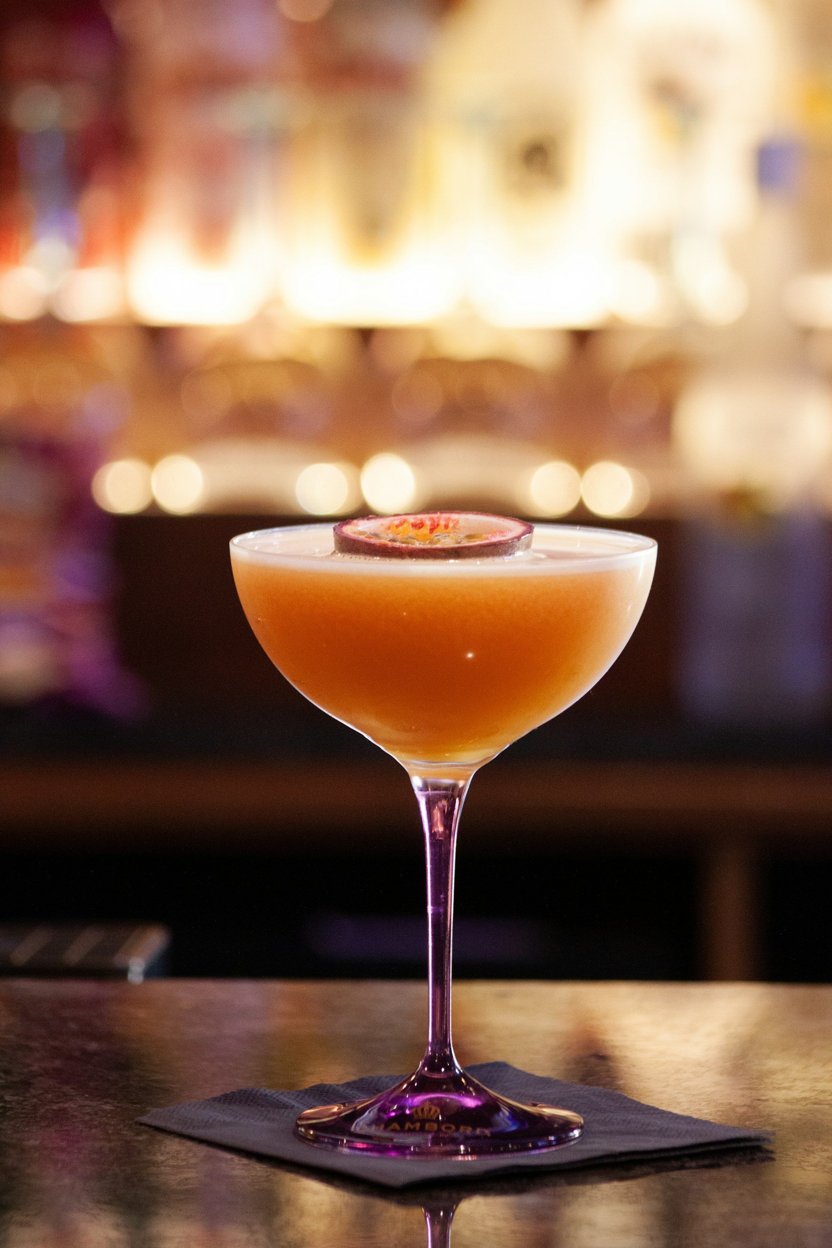 A cocktail in a coupe glass with a decorative slice of fruit on top, placed on a napkin on a bar counter with blurred warm lighting in the background.