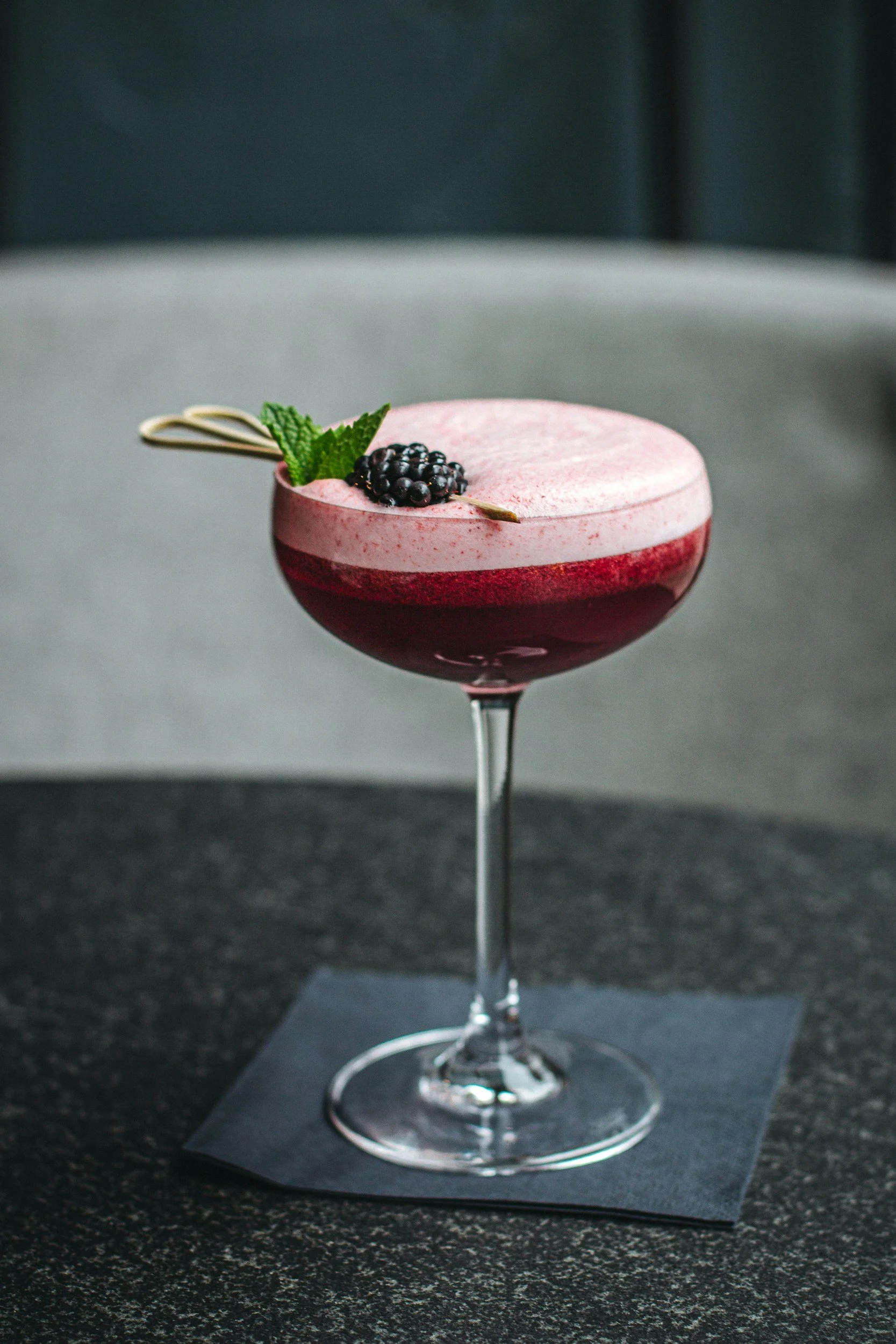 A pink cocktail with foam, garnished with a blackberry, mint leaves, and a decorative white chocolate stick, in a coupe glass on a dark napkin.