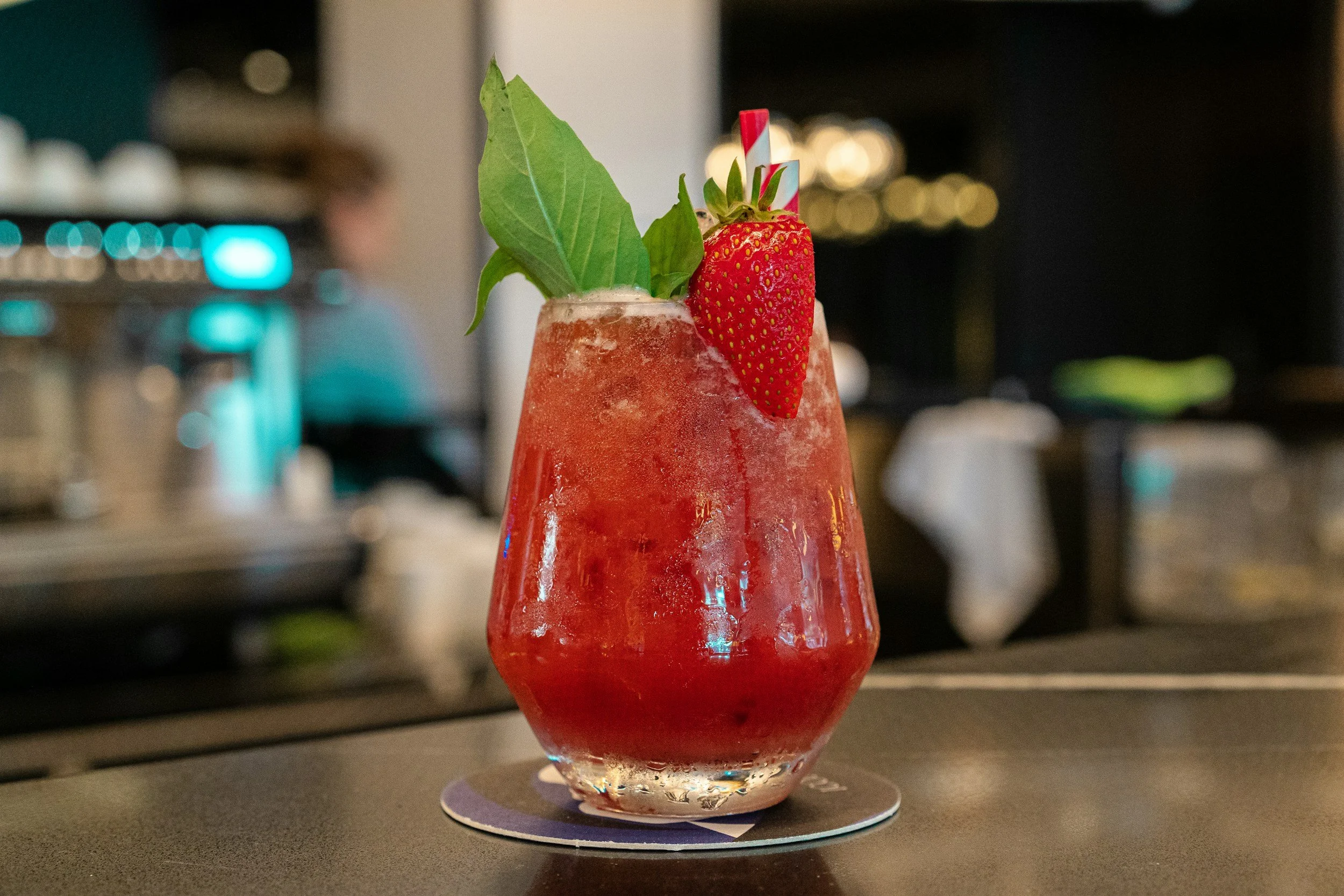 A tall glass filled with a red strawberry drink, garnished with a whole strawberry, a green leafy herb, and a striped straw, placed on a round paper coaster on a dark countertop.