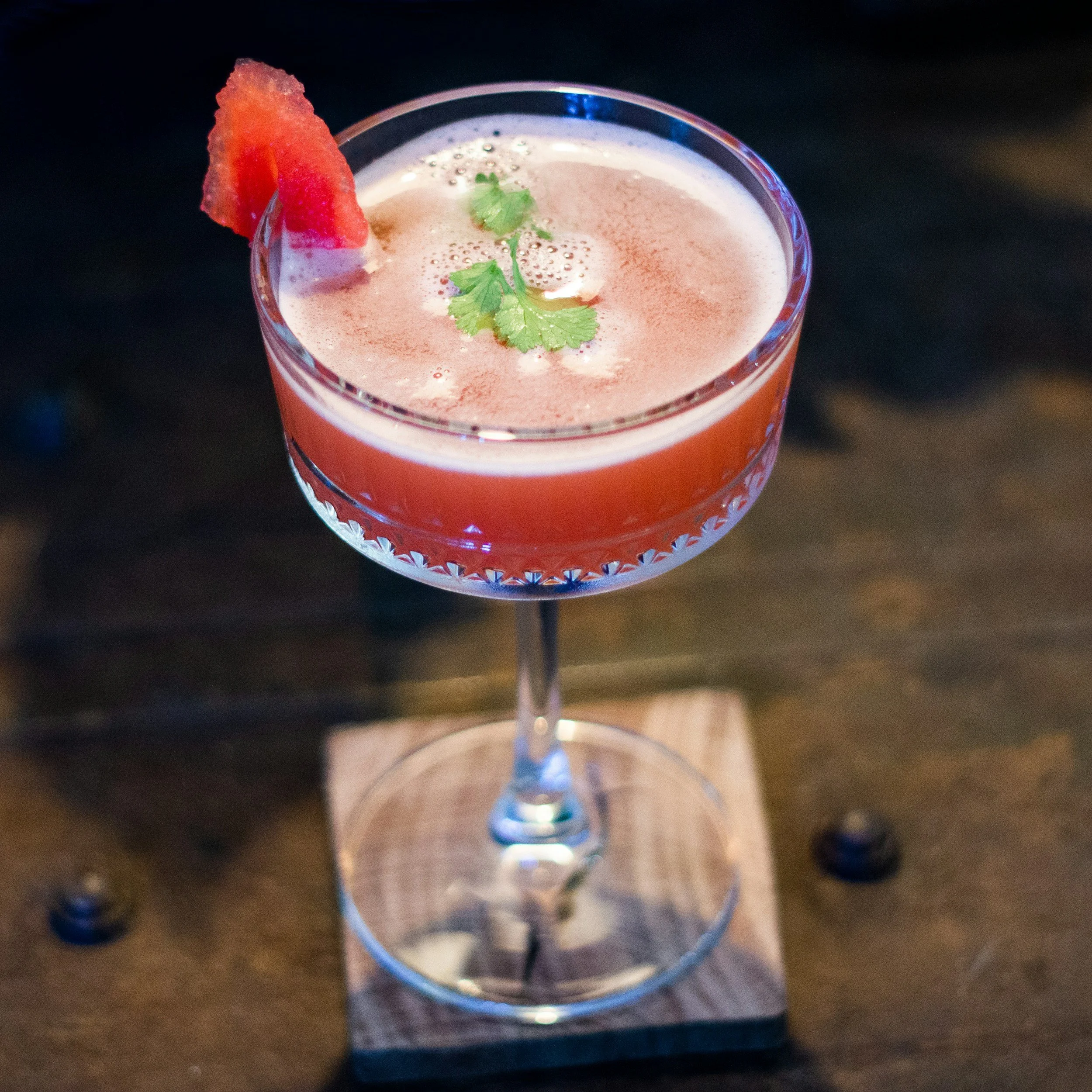 A cocktail drinks with a pinkish color in a clear glass tumbler, garnished with a slice of watermelon and a sprig of cilantro, with a foam layer on top.
