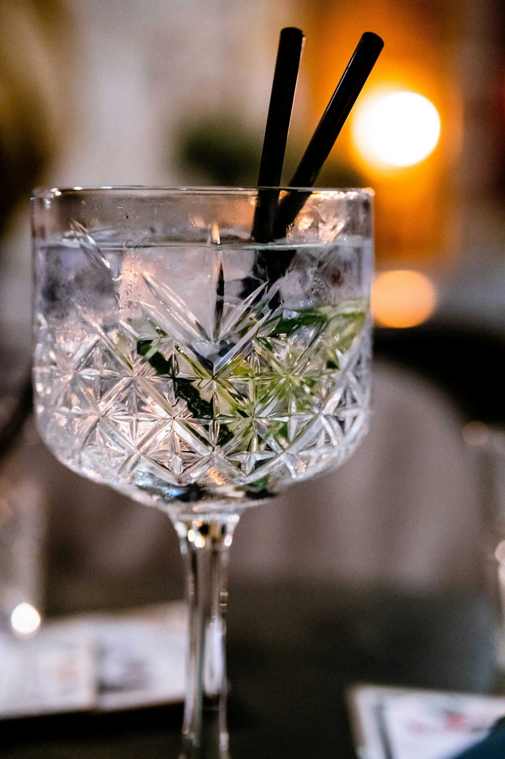 Close-up of a glass of clear beverage with ice and mint leaves, two black drinking straws, decorated with an intricate cut pattern, set against warm blurred background with bokeh lights.