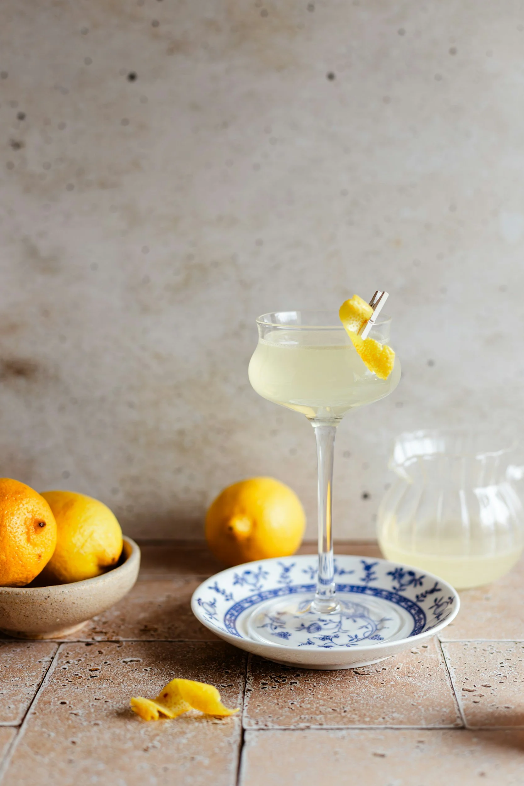 A glass of lemon cocktail with a lemon peel garnish on a blue and white patterned plate. A bowl of lemons and a glass pitcher are in the background. The surface is a tiled countertop, and the background is a textured gray wall.