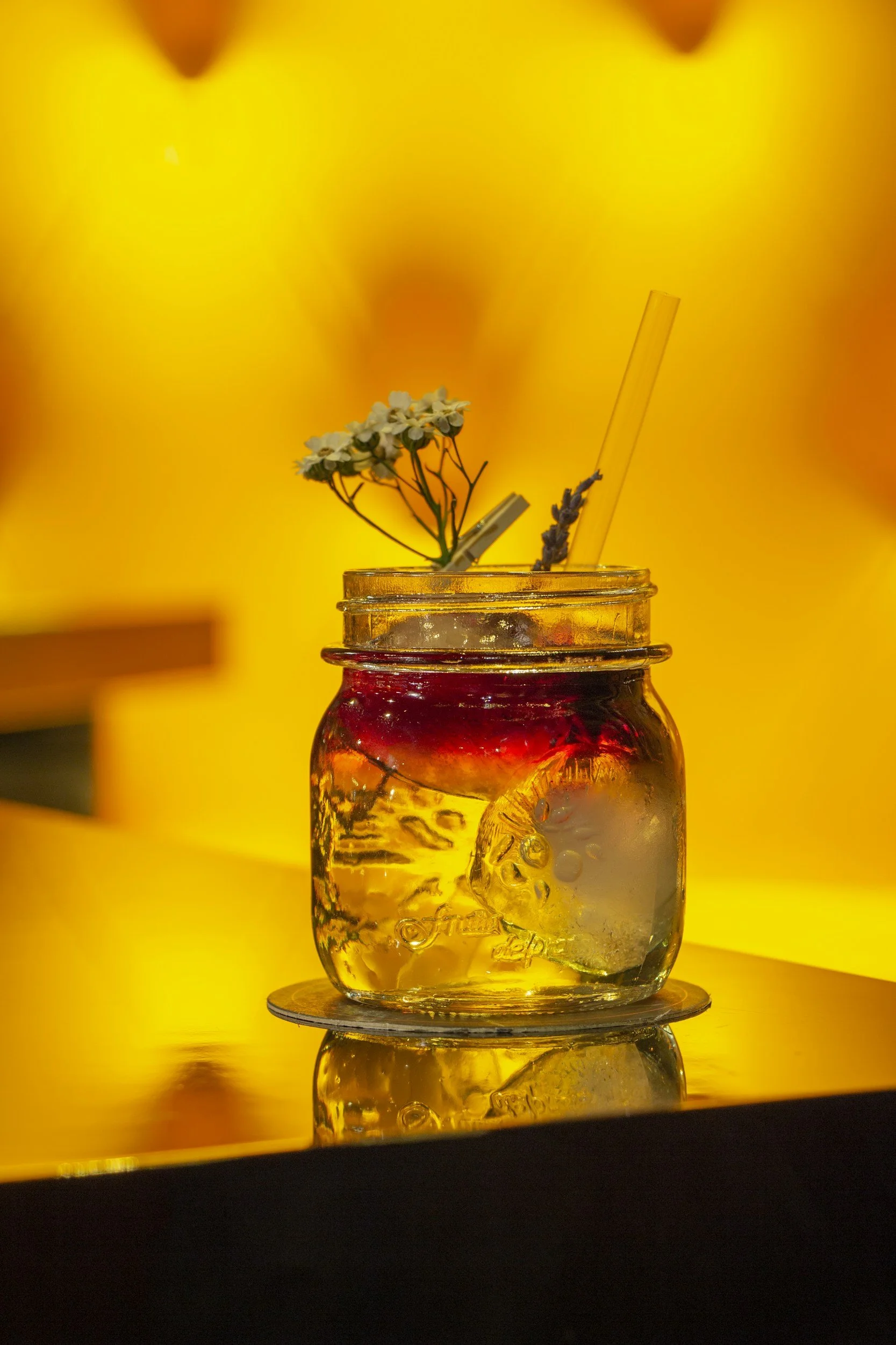 A mason jar filled with a yellow-colored beverage with ice cubes inside. There is a small bunch of white flowers and a lavender sprig in the jar. A clear straw is inserted into the drink. The jar is placed on a round coaster on a reflective surface, 