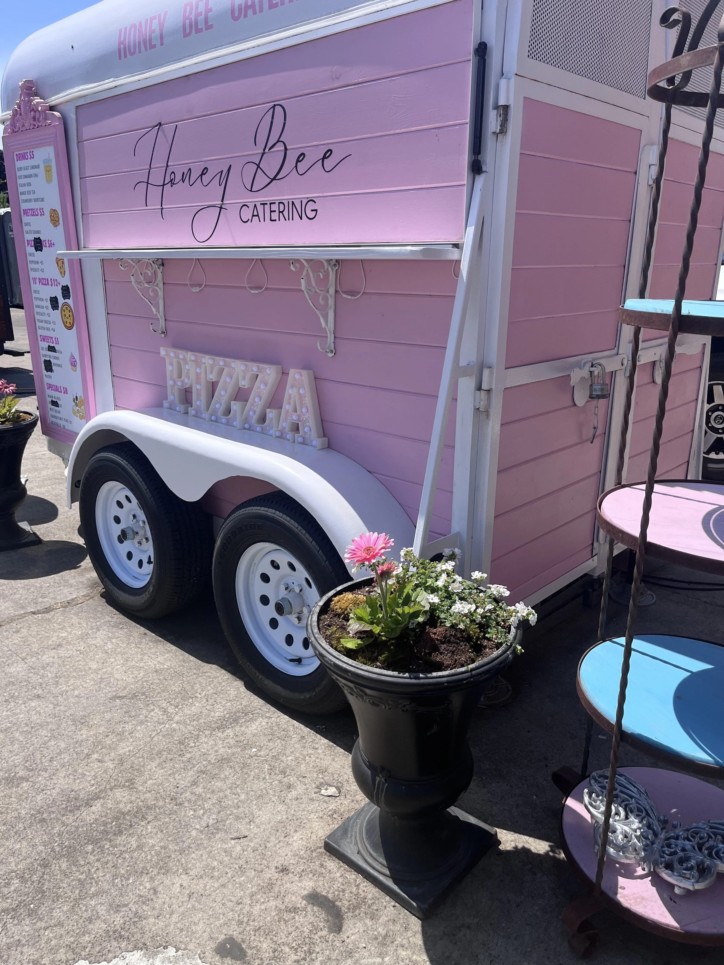 A pink mobile catering trailer with the words 'Honey Bee Catering' on the side, with a menu board listing drinks, pretzels, pizza, and sweets, and decorative flowers in black planters and outdoor tables around it.