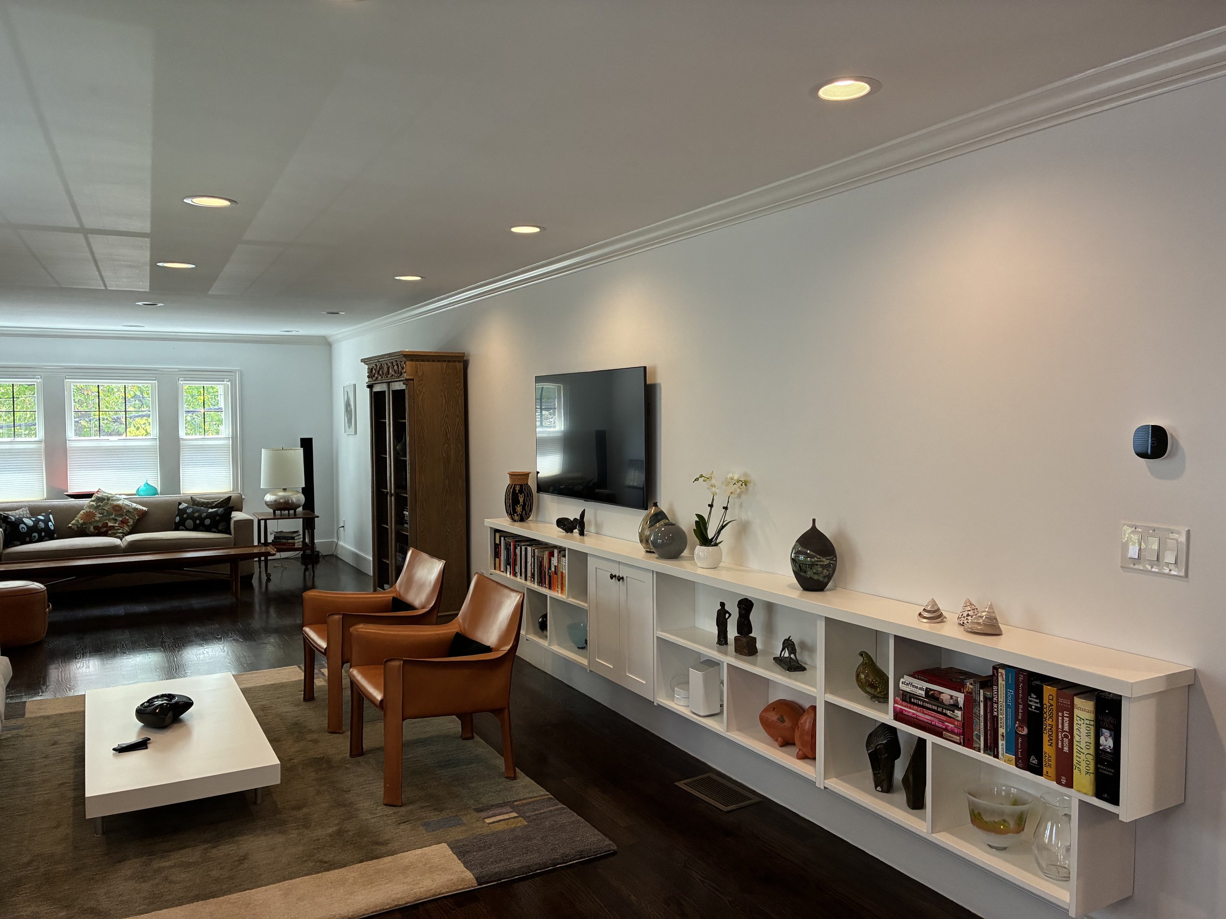 Living room with white walls, wooden furniture, a flat-screen TV, white bookshelf with decorative items and books, black ceiling speaker, brown chairs, white coffee table with a remote, and windows with blinds.