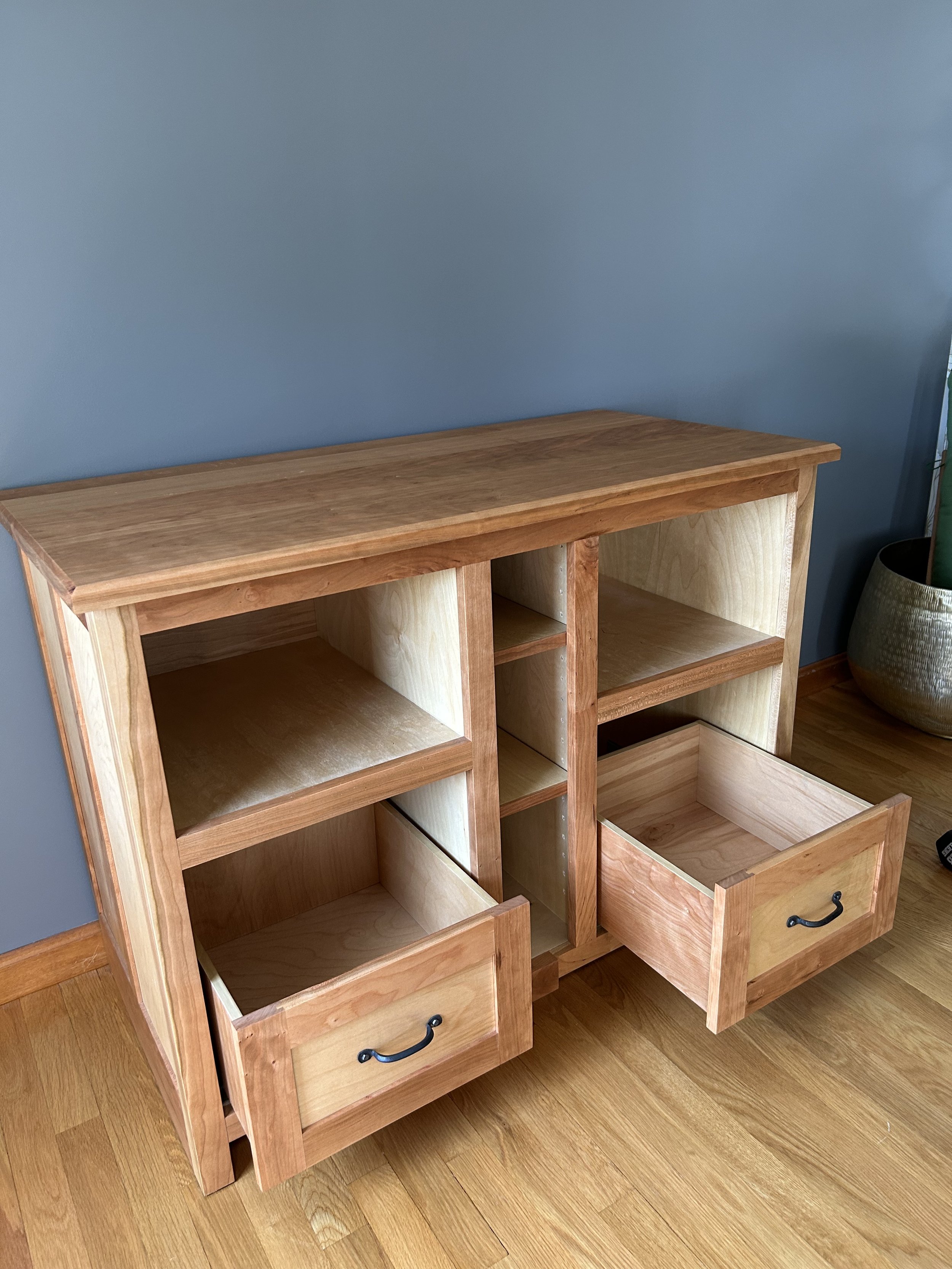 unfinished wooden bookshelf with open shelves and a drawer, placed against a blue wall with a potted plant to the right.