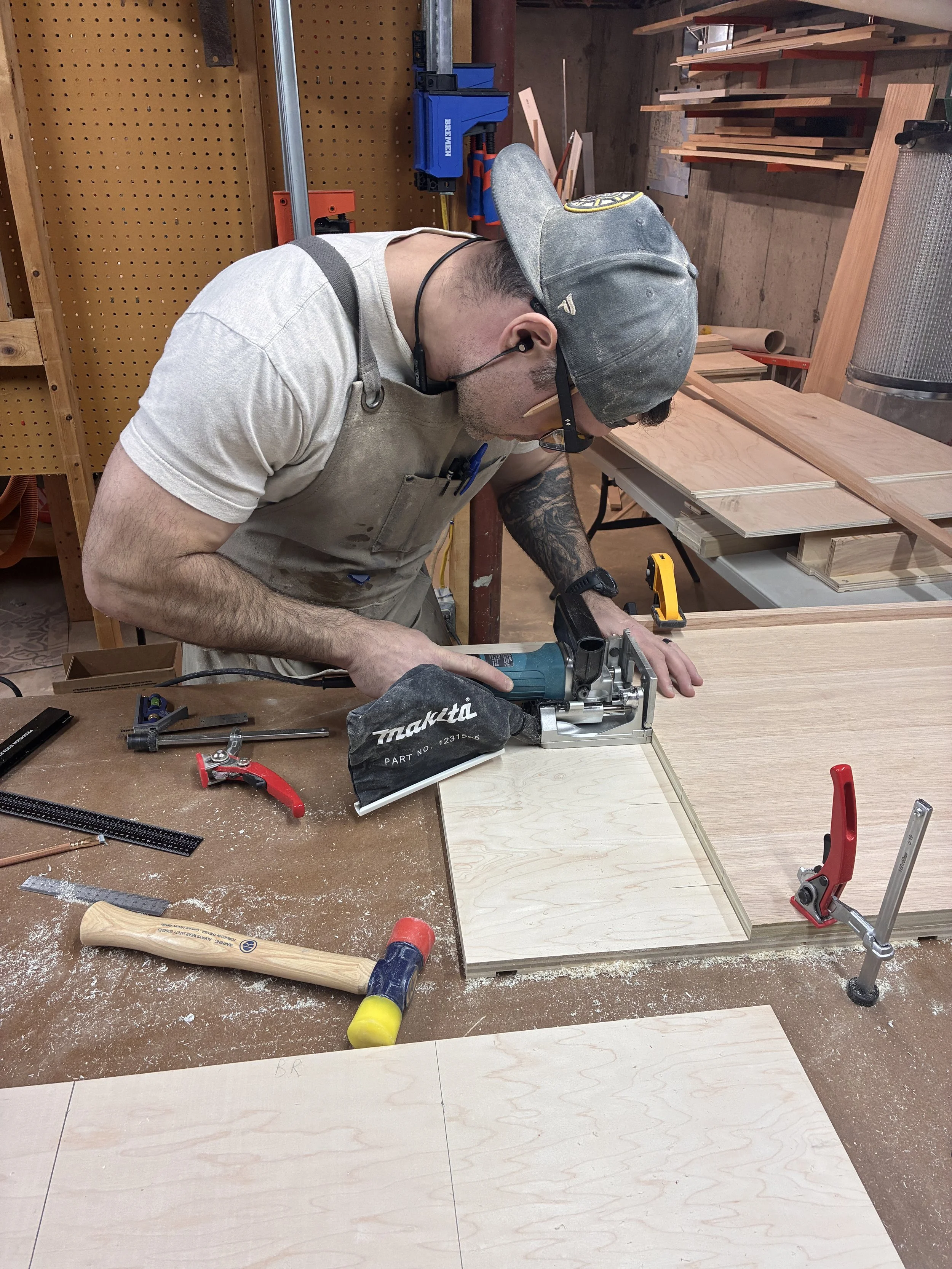 A man in a gray cap, glasses, and a beige apron is working in a woodworking shop. He is using a power tool on a piece of wood. The workshop has various hand tools, small wood pieces, clamps, and a pegboard with hooks for tools.