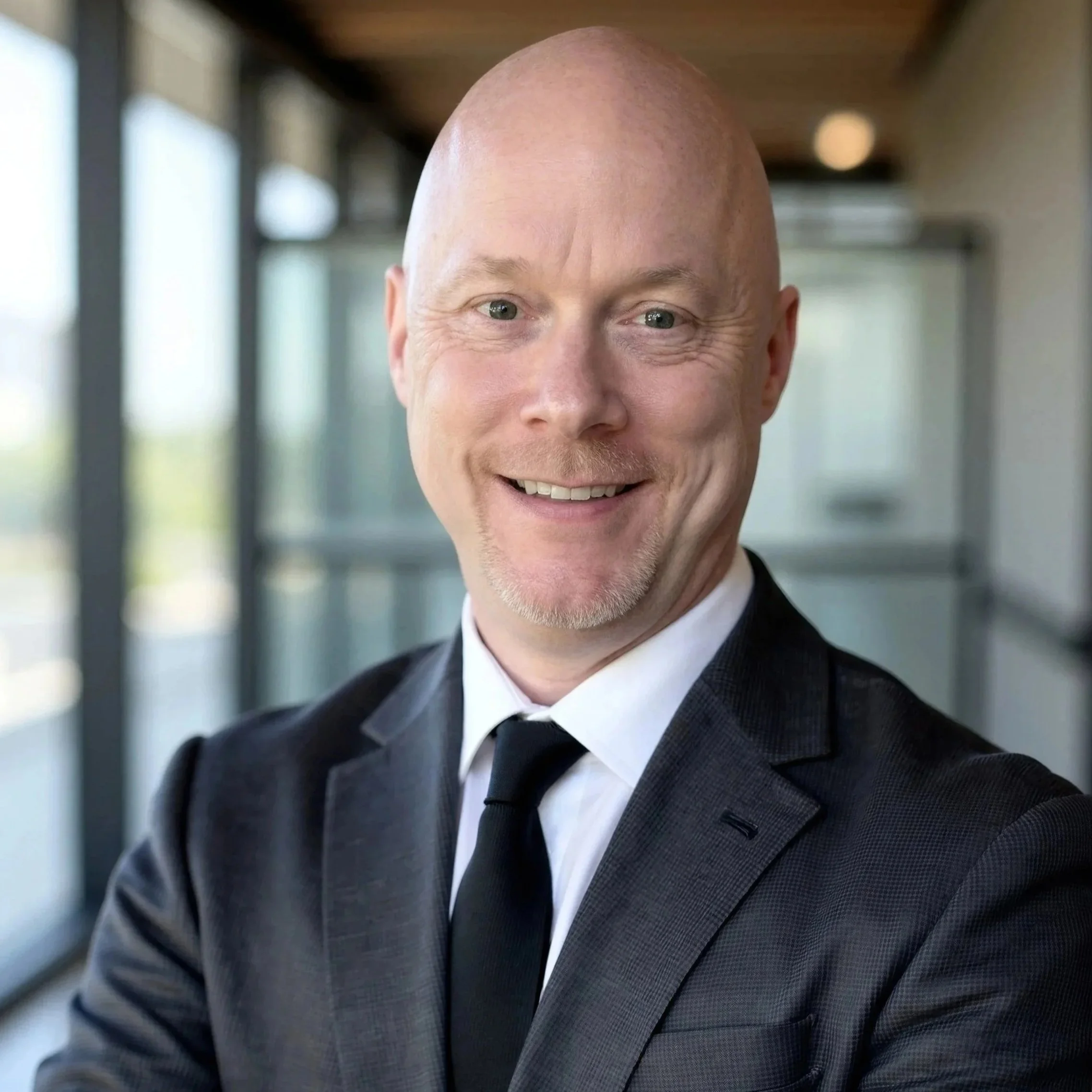 Close-up of a smiling man with a bald head, wearing a dark suit and white shirt, standing in a modern building with large windows.