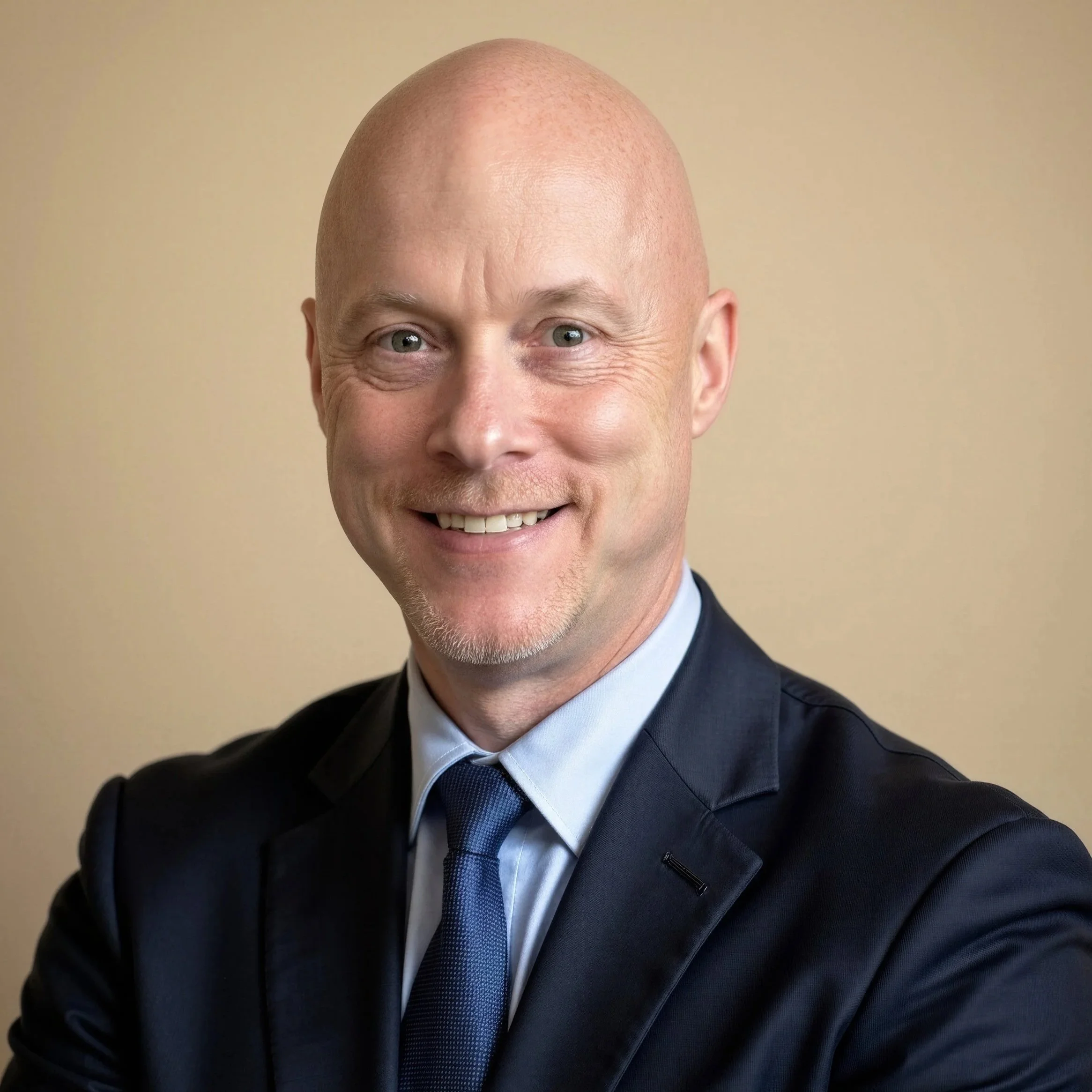 Close-up of a smiling man with a shaved head, wearing a dark suit, white shirt, and blue tie, against a plain beige background.
