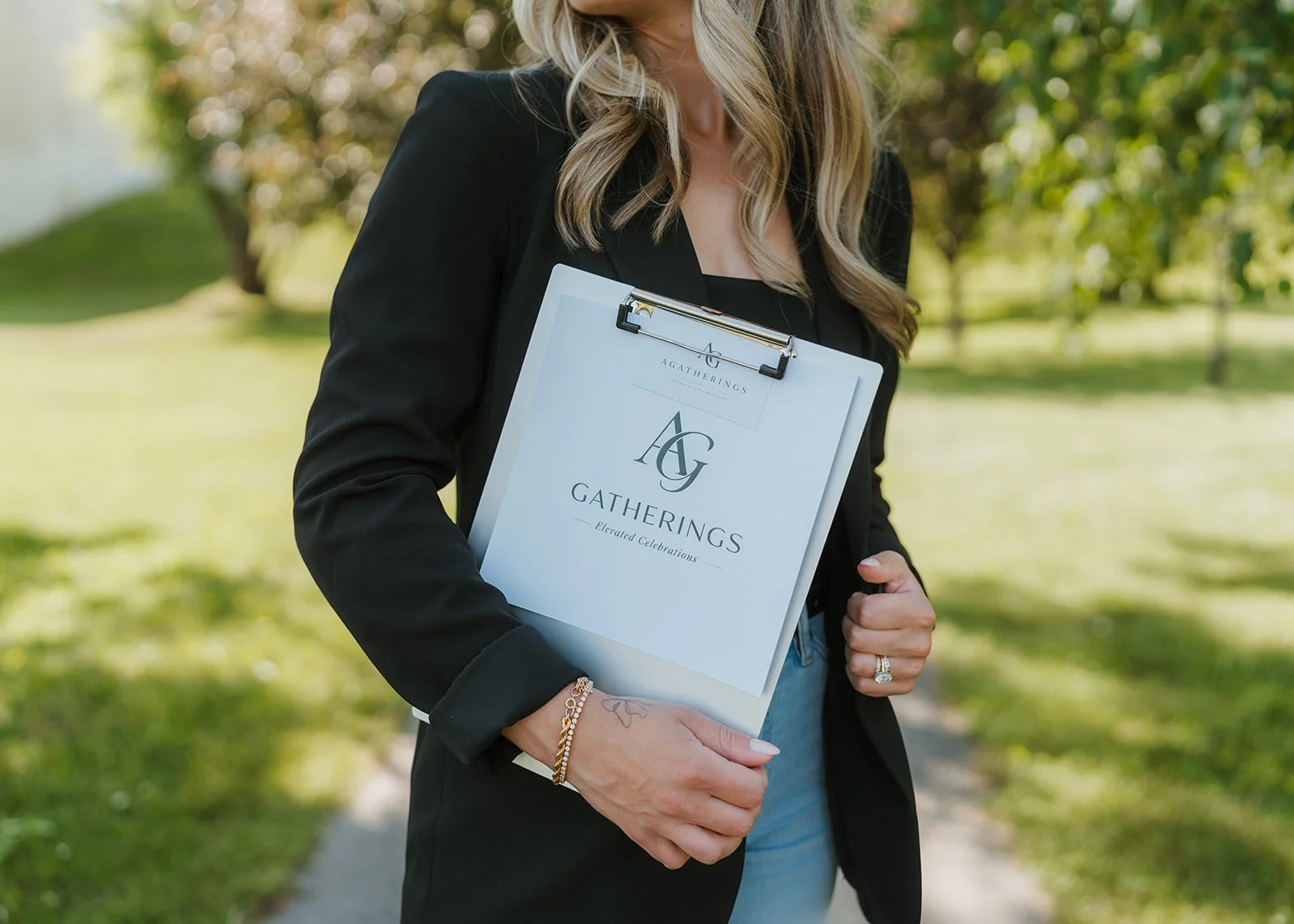 A woman holding a clipboard with a paper that reads 'Gatherings' and an 'AG' logo, walking outdoors in a park with green trees and grass.