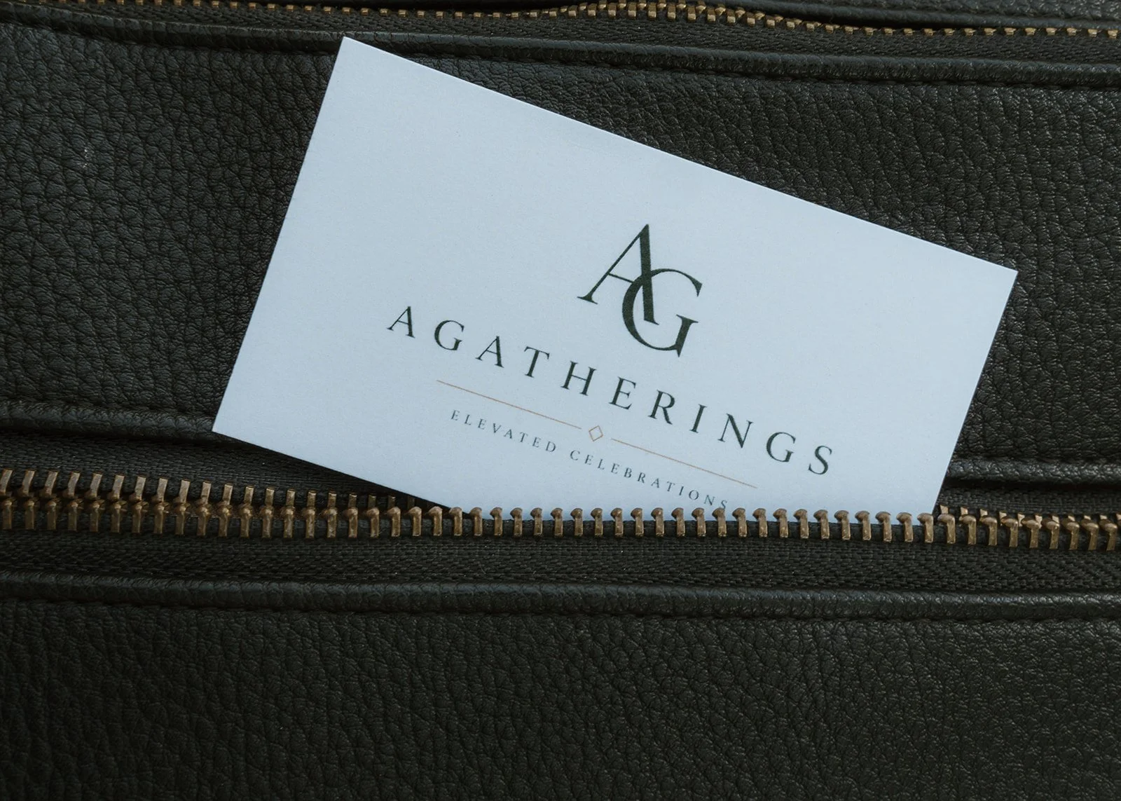 A business card with the initials 'A G' and the name 'Agatherings,' along with the tagline 'Elevated Celebrations,' lying on a textured black surface with a zipper.