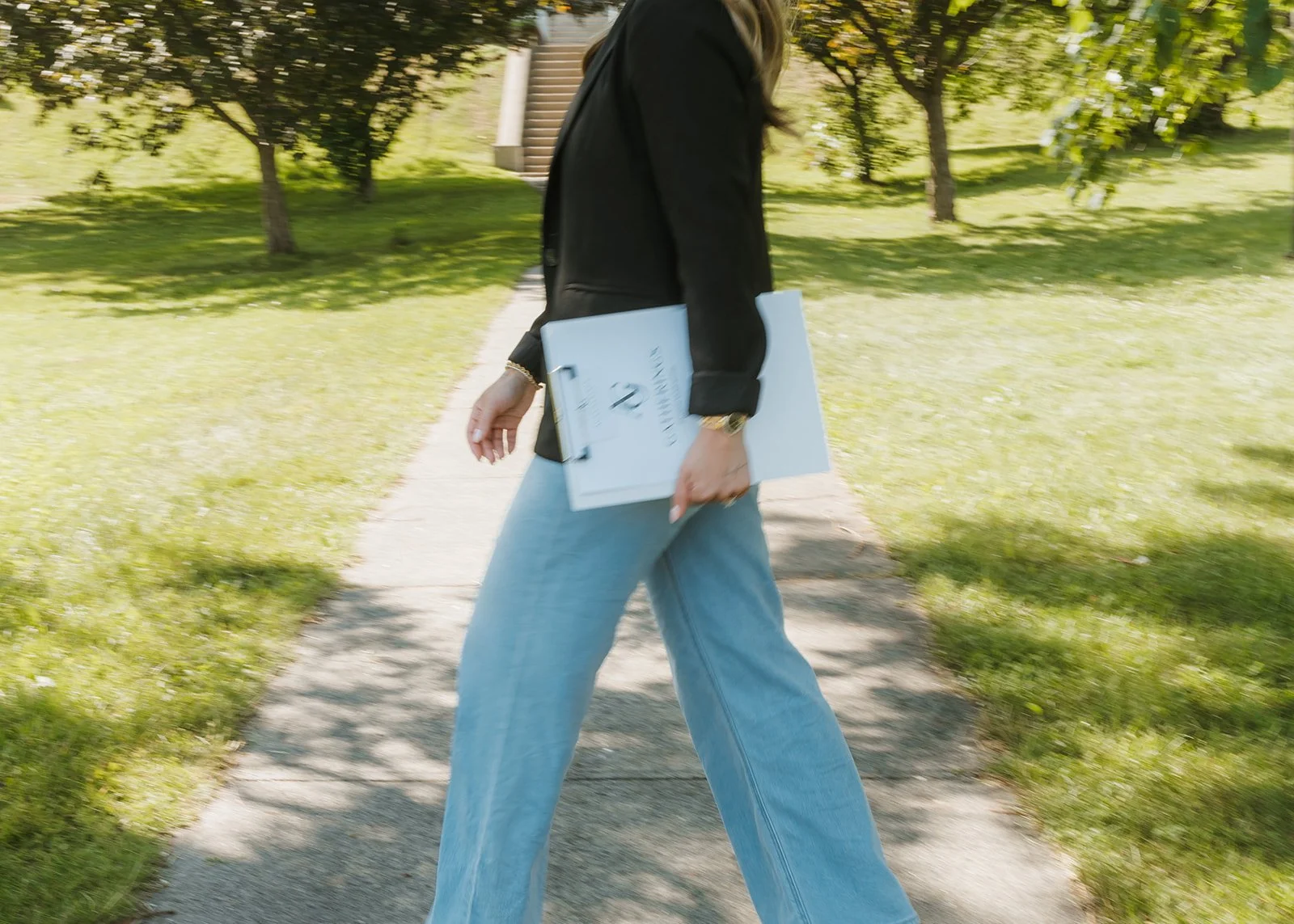 A woman walking on a sidewalk in a park, carrying a clipboard or folder, with trees and grass in the background.
