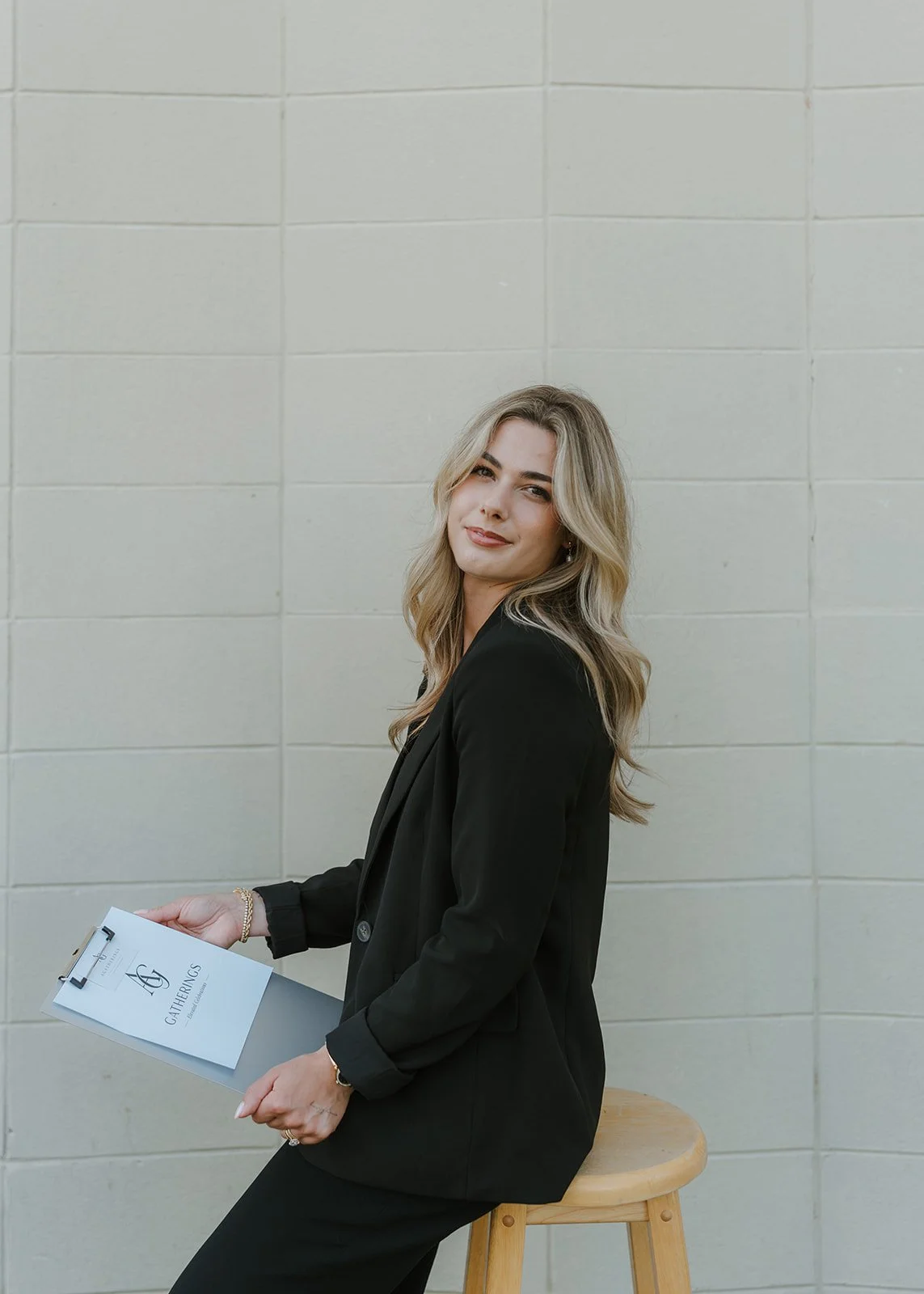 A woman with blonde hair wearing a black blazer and pants, sitting on a wooden stool, holding a clipboard with a paper titled 'GATHERINGS,' against a plain tiled wall.