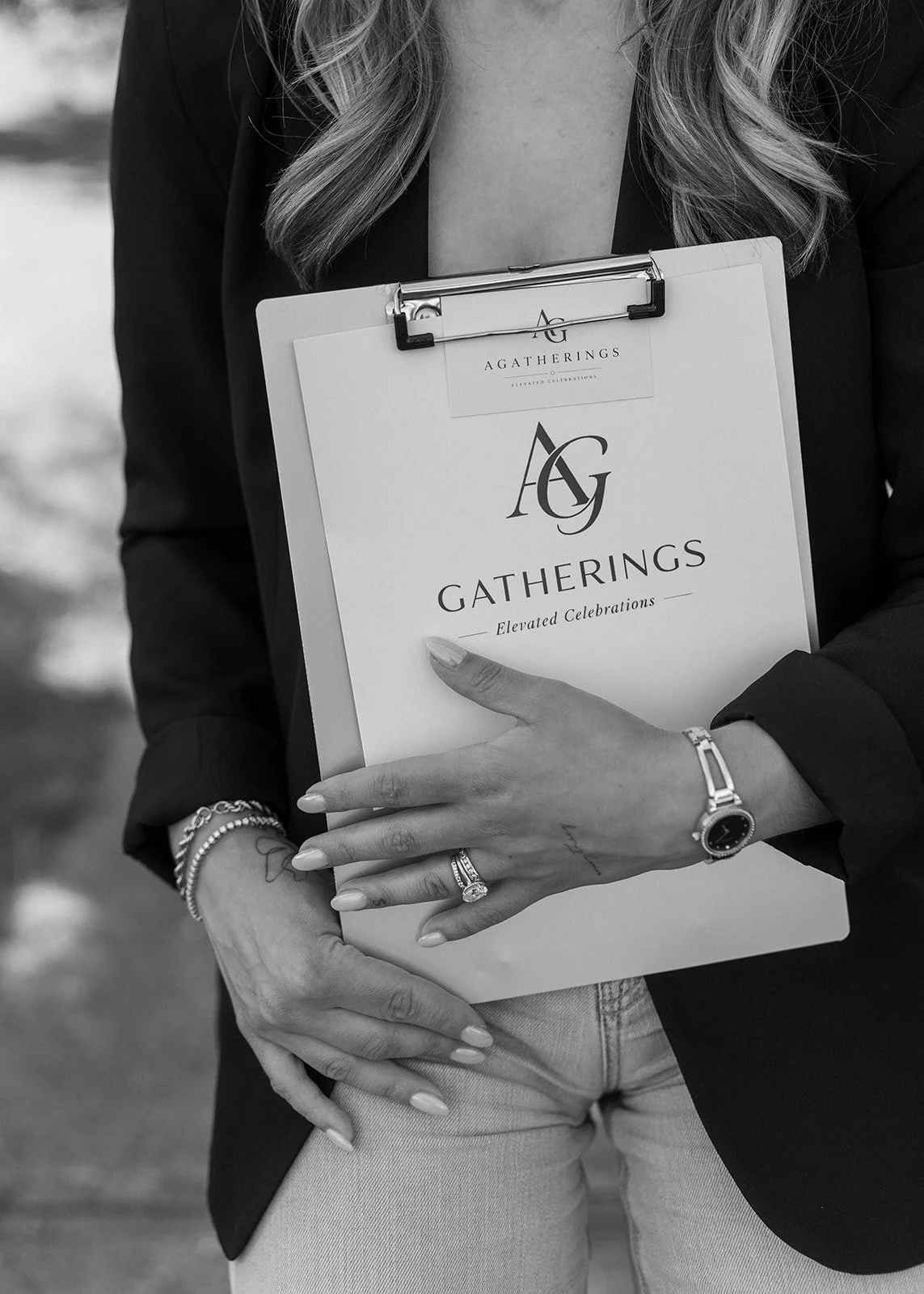 Black and white photo of a woman holding a clipboard with the logo and name 'Gatherings Elevated Celebrations'. She is wearing a watch and rings on her left hand, a bracelet, and a dark blazer.