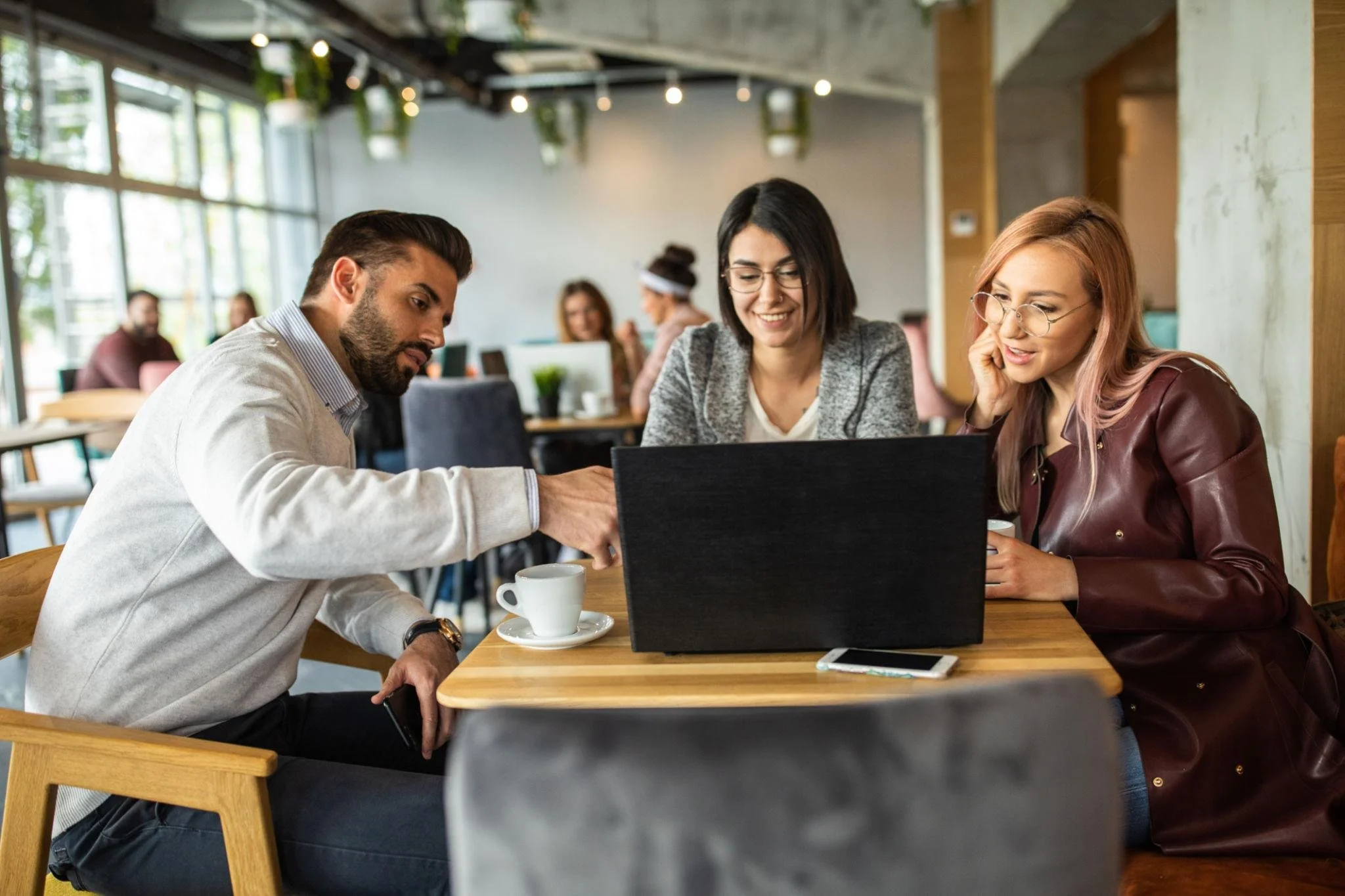 Tres personas trabajan juntas en una computadora portátil en una cafetería moderna. Dos mujeres y un hombre parecen concentrados en el trabajo y están sentados alrededor de una mesa de madera. Hay tazas blancas en la mesa y otros clientes en el fondo.