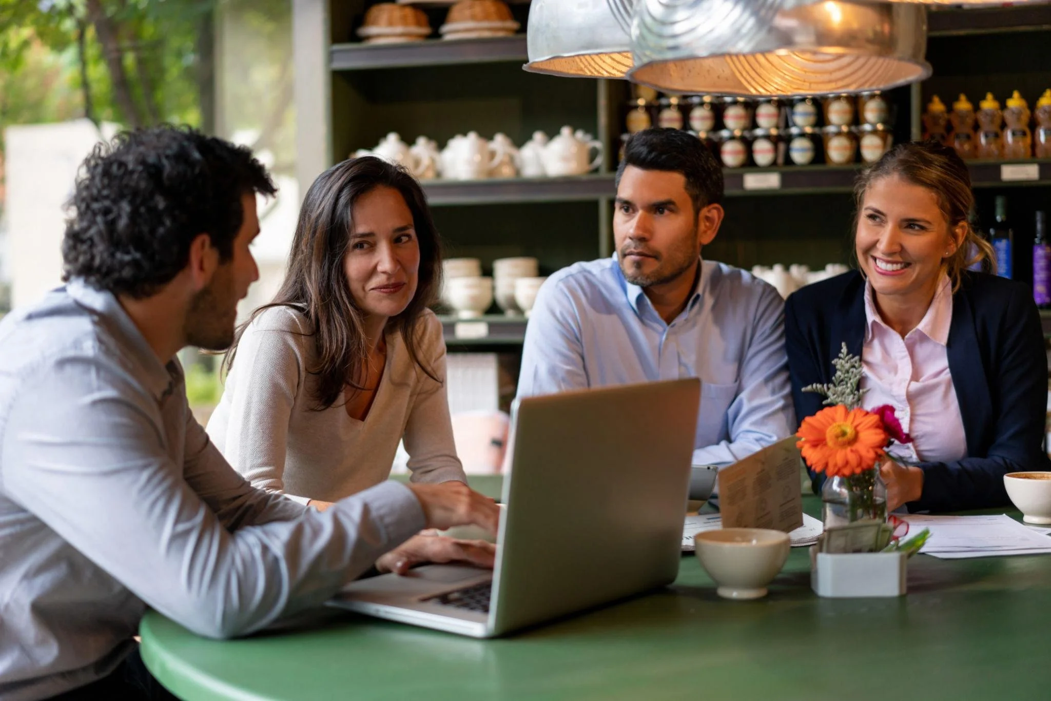 Grupo de adultos en una reunión de negocios en una cafetería, con una laptop en la mesa y decoraciones con flores