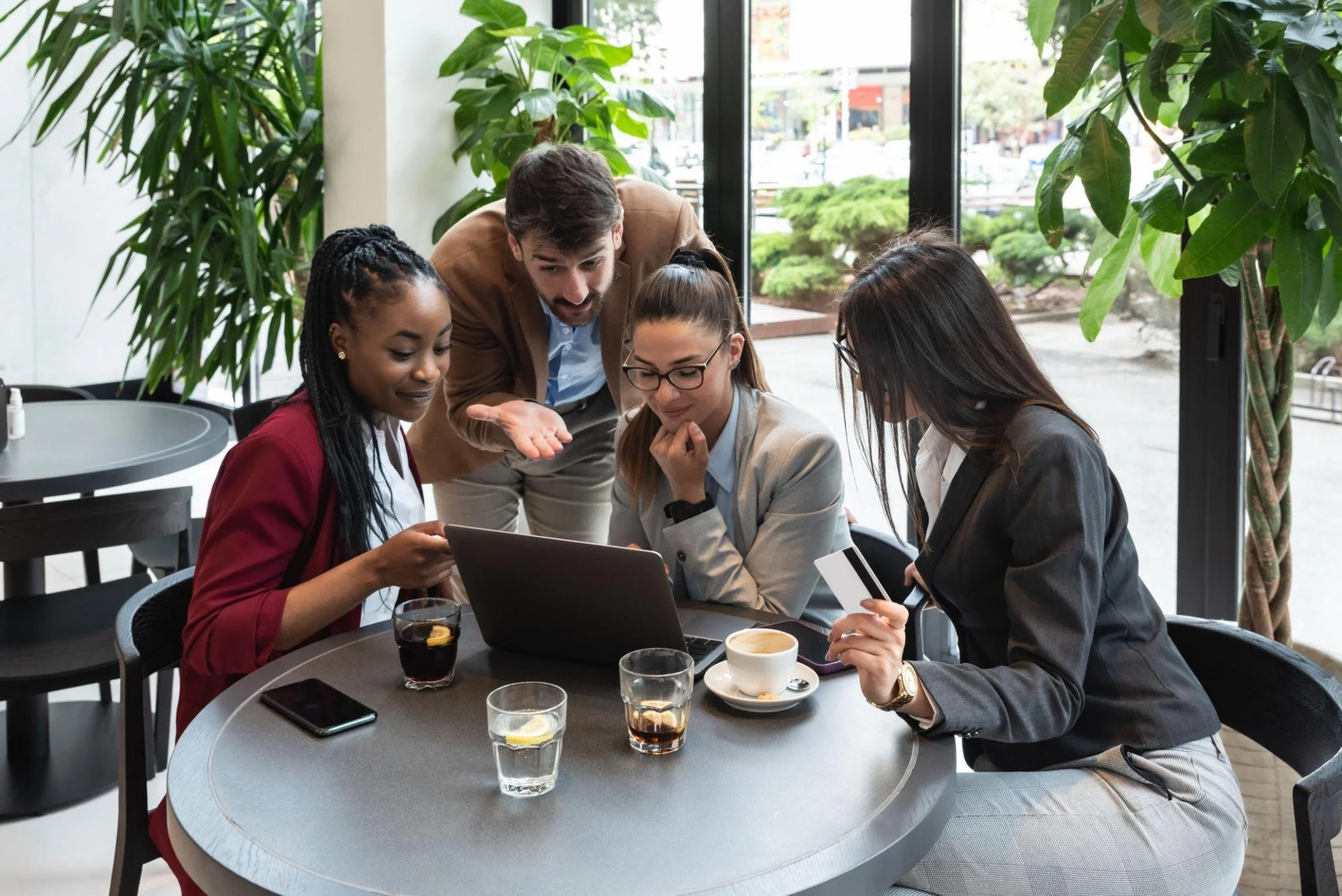 Grupo de cinco personas en una cafetería, observando una pantalla de computadora portátil, con bebidas en la mesa y plantas verdes alrededor.