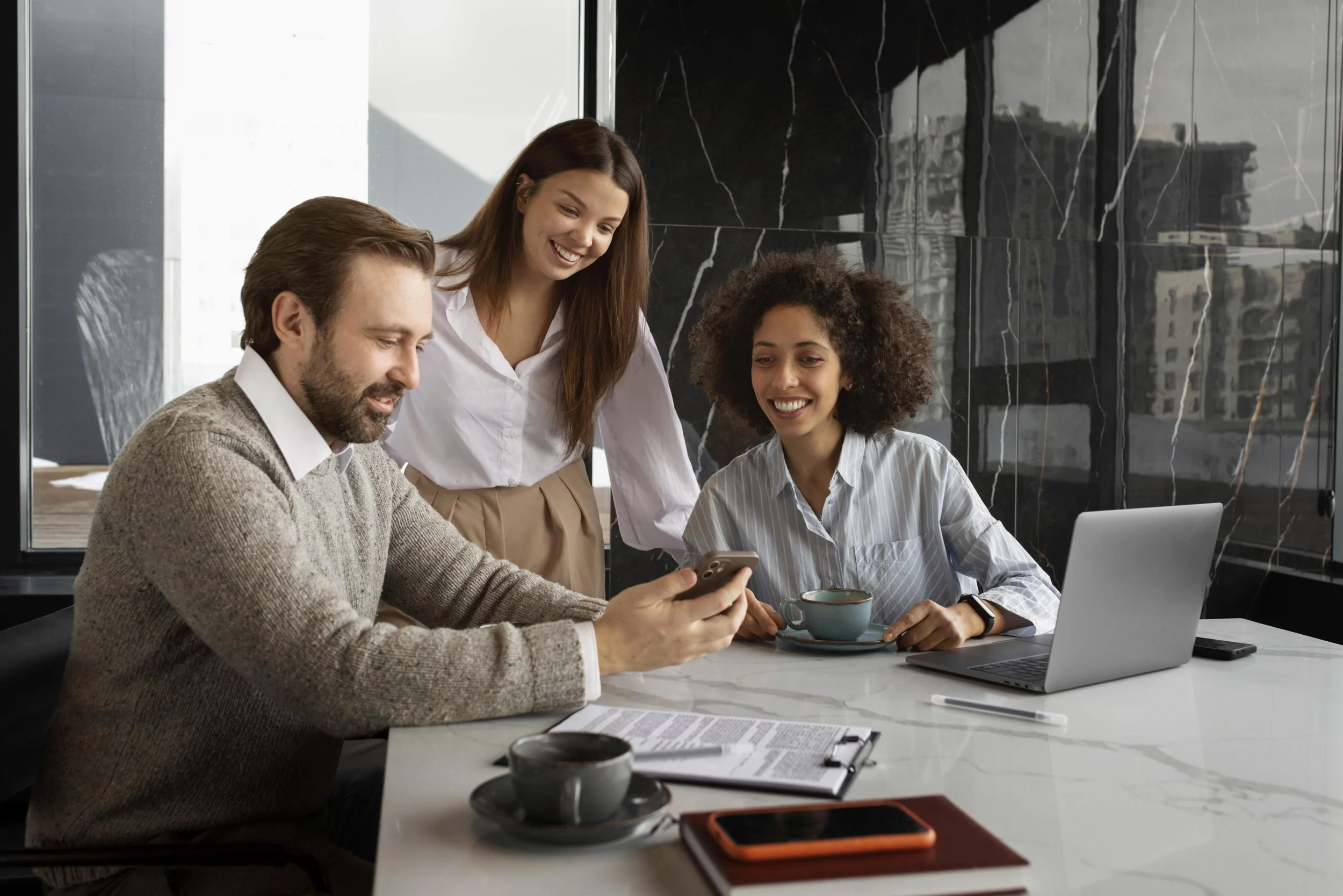 Tres personas en una reunión de trabajo en una oficina moderna, una mujer y un hombre viendo una pantalla y otra mujer observando y sonriendo.