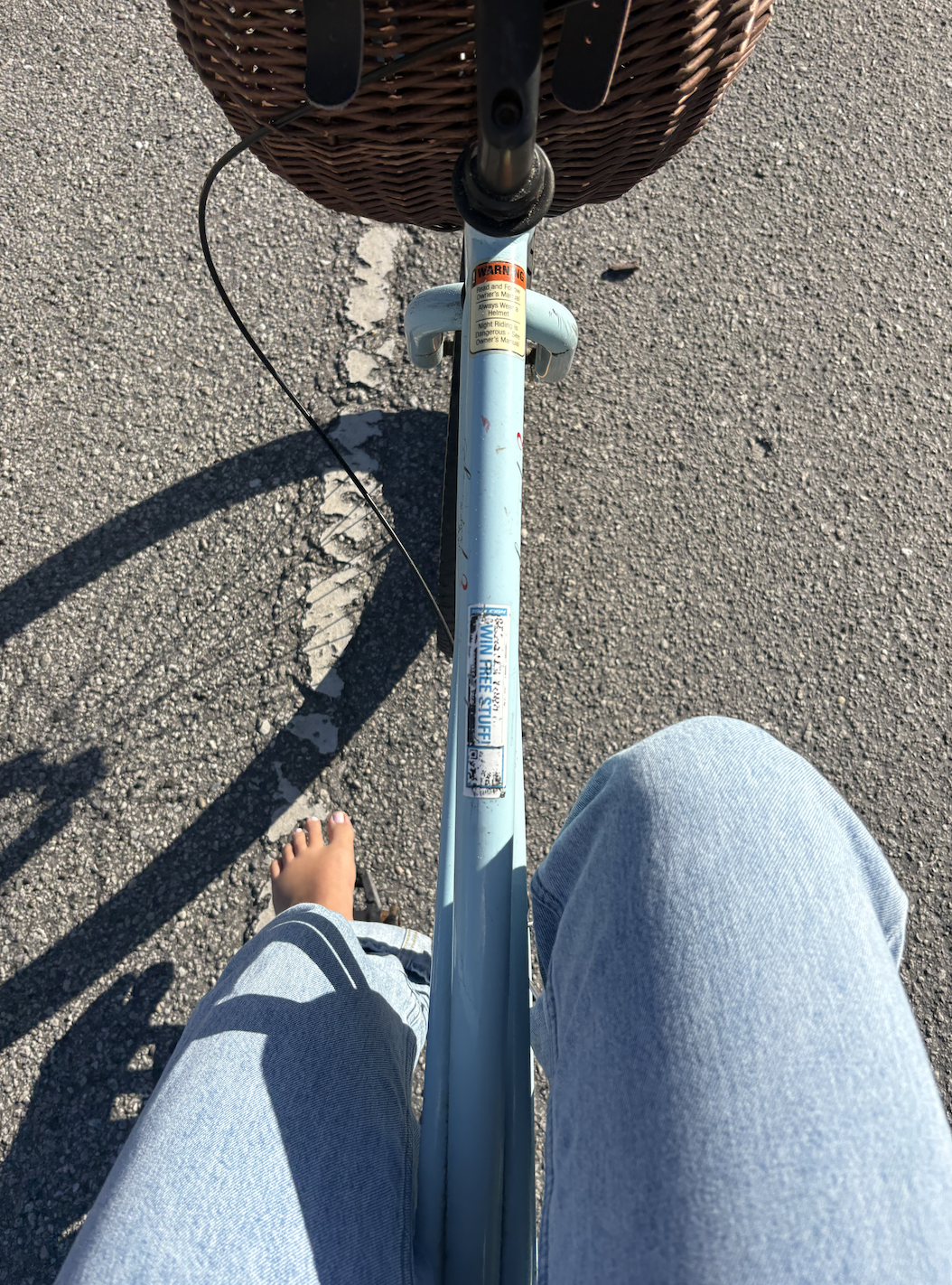 First-person view of a person riding a bicycle on a paved road, barefoot with jeans, casting a shadow on the asphalt, with a brown wicker basket attached to the front of the bike.