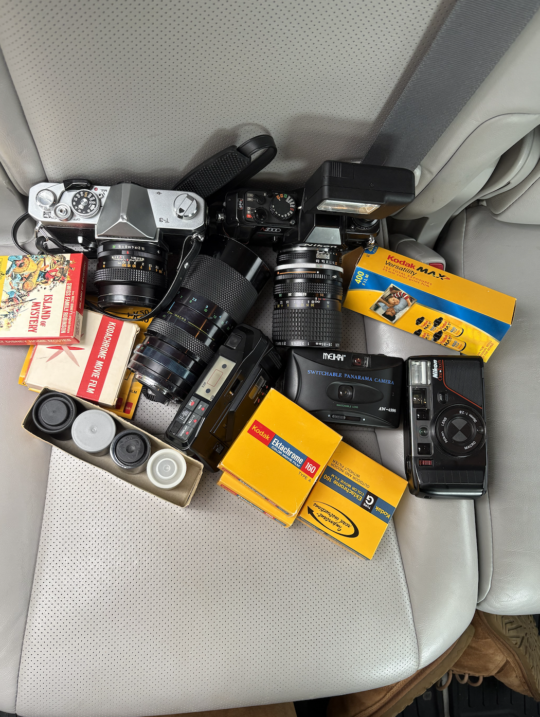 Collection of vintage cameras, film rolls, and photography accessories on a car seat.
