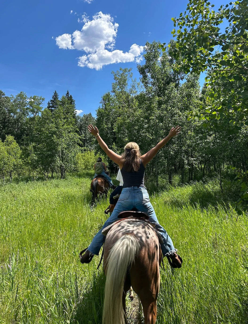 A woman riding a horse with arms raised, leading a group of people riding horses through a green forested area on a sunny day.