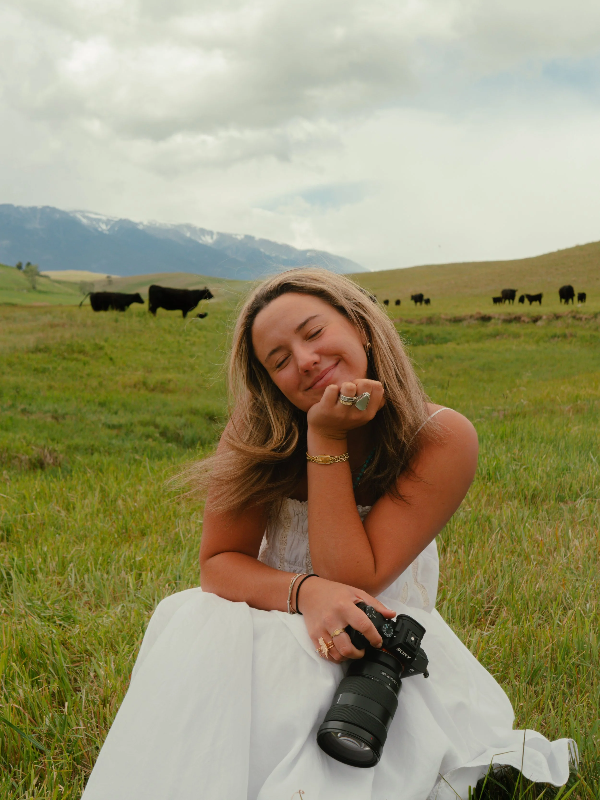 A young woman with long hair smiling and resting her chin on her hand, sitting on a grassy field with rolling hills, cows, and mountains in the background, holding a camera in her lap.