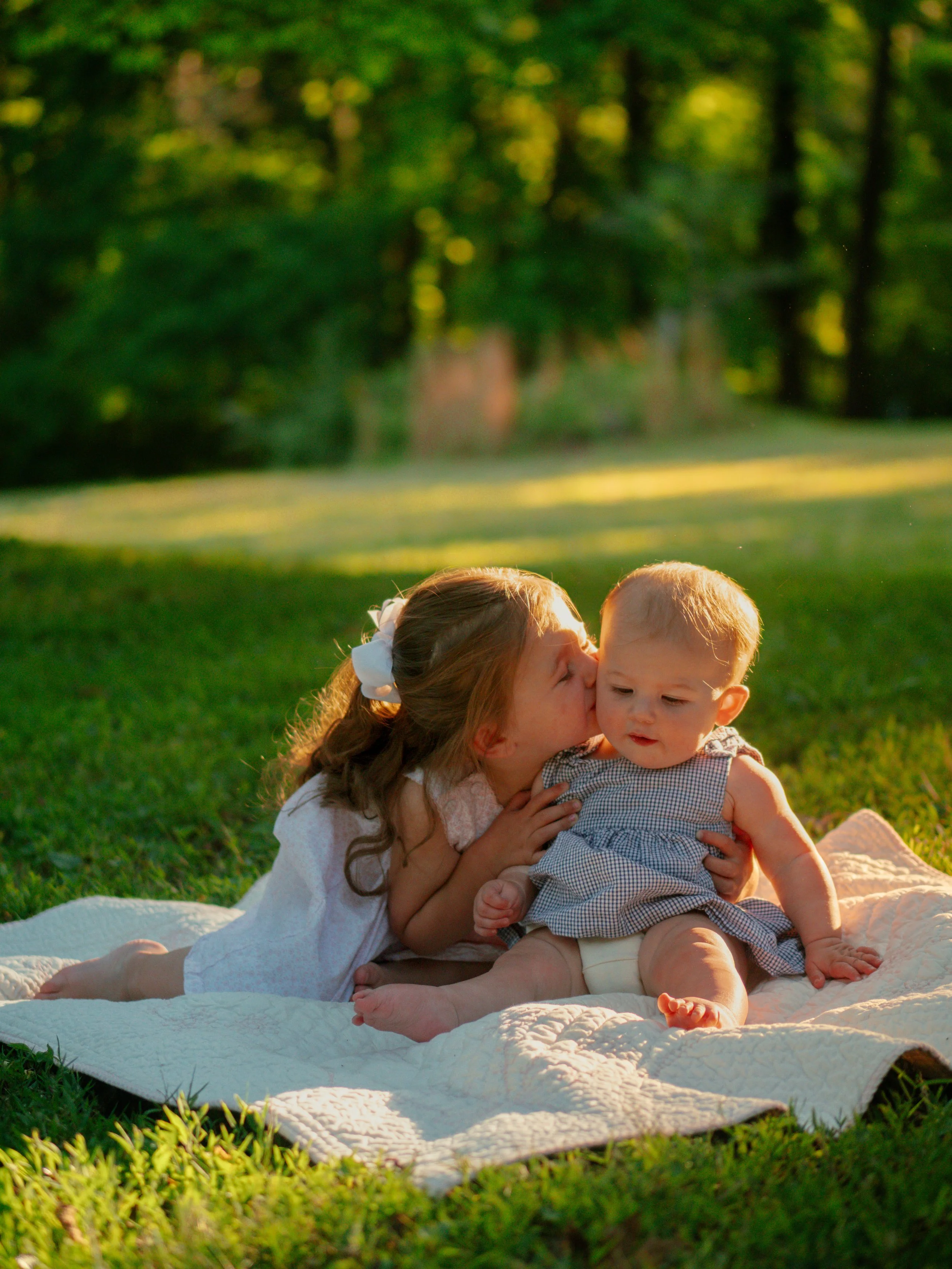A woman and a young child sit on a cream quilt outdoors in a park, with lush green grass and trees in the background, during what appears to be late afternoon or early evening sunlight.