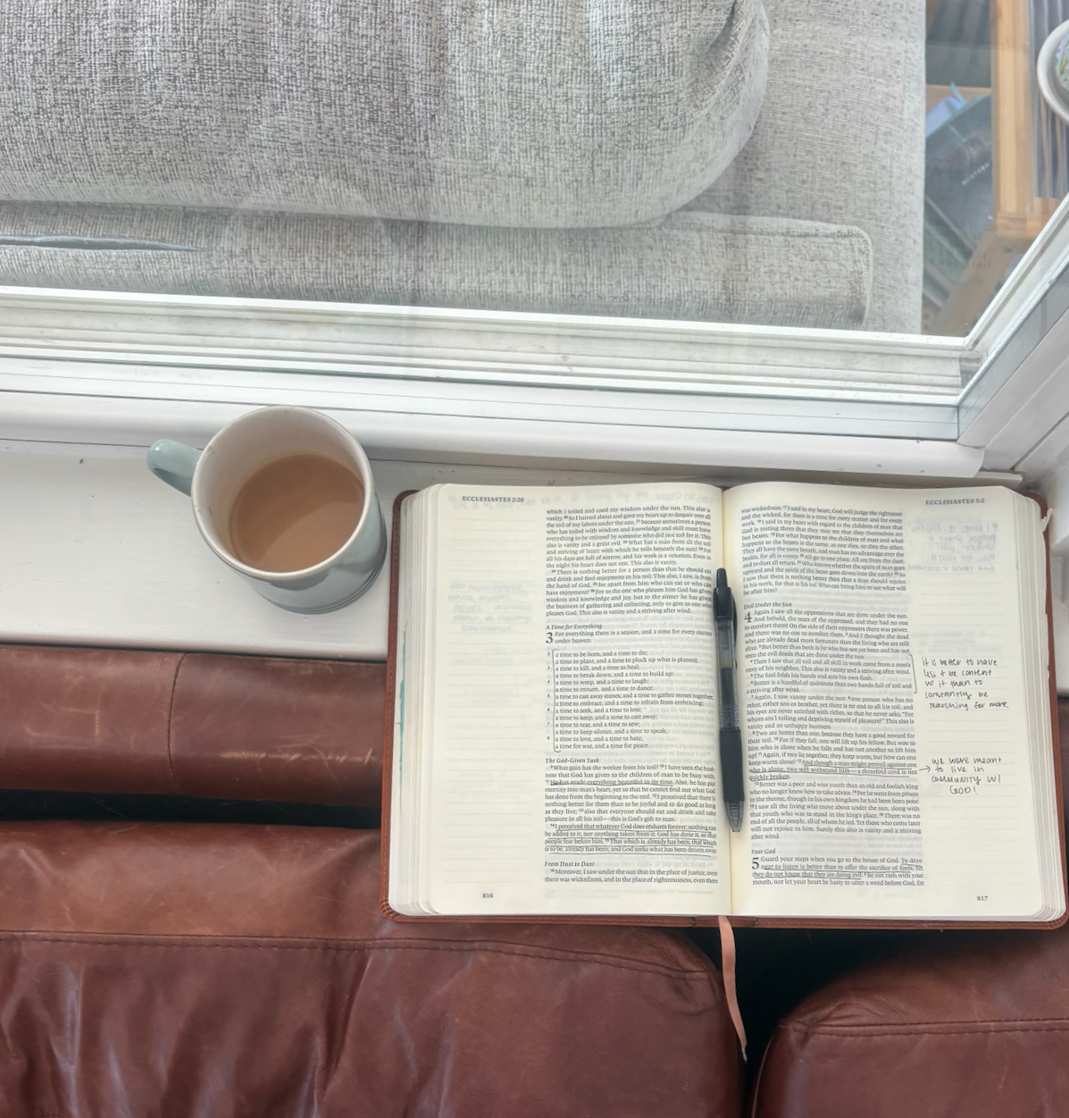 A book, a mug of coffee, and a pen placed on an open Bible, viewed through a window with a couch and a wooden table in the background.