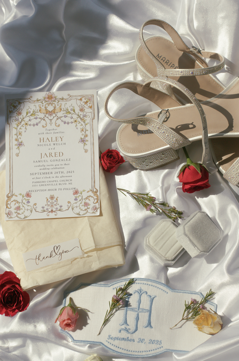 Wedding invitation, red and pink roses, sprigs of greenery, a pair of silver rhinestone heels, a floral monogram napkin with the date September 20, 2025, and a small thank you note, all arranged on satin fabric.