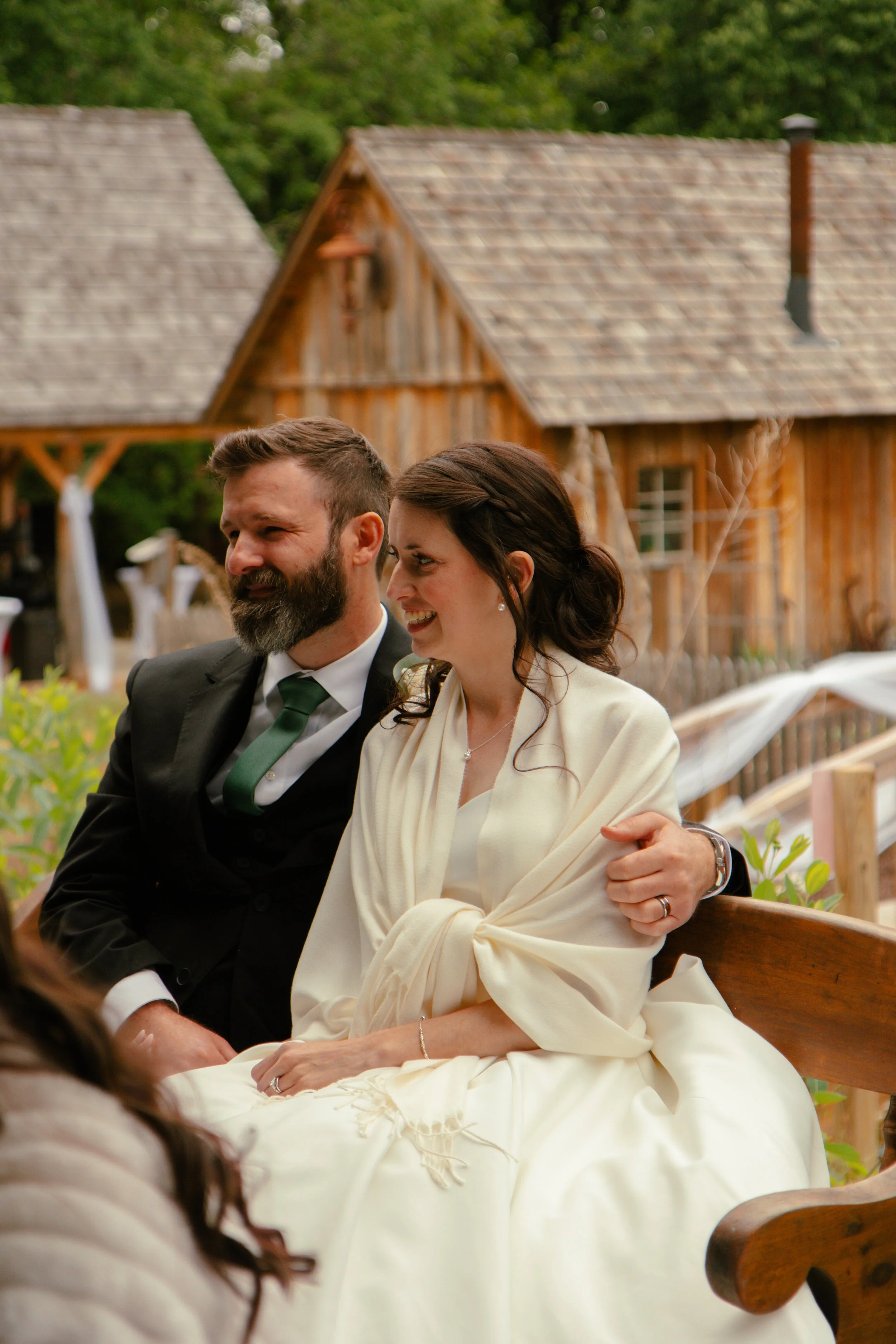 A man with a beard and a woman with dark hair sitting close together on a wooden bench at an outdoor event. The man is wearing a black suit with a white shirt and green tie, and the woman is wearing a white shawl over a white dress with her hair styled in a braid. Wooden buildings and greenery are in the background.