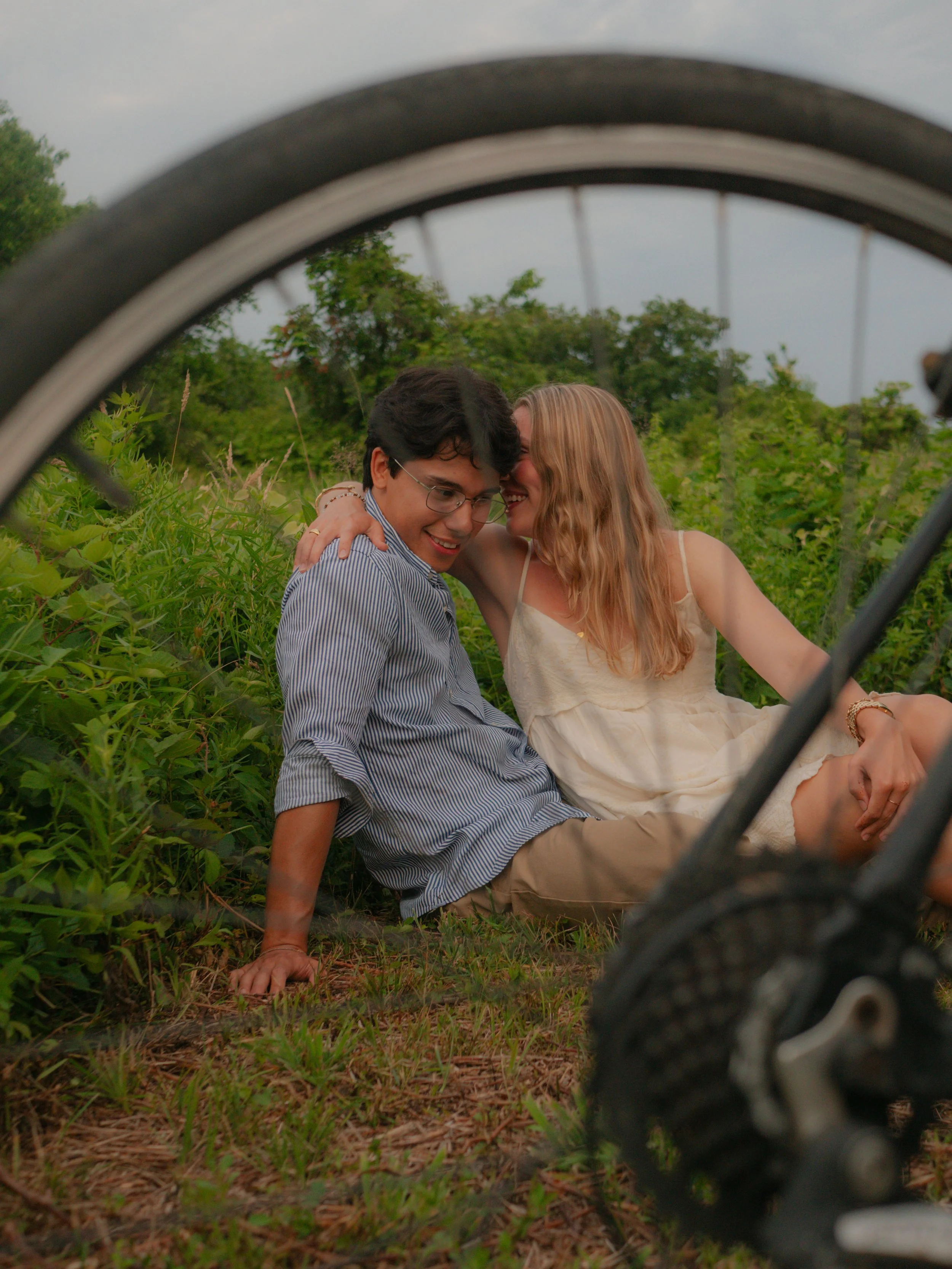 A couple sitting in a grassy field, smiling and leaning close to each other, viewed through the tire of a bicycle in the foreground.