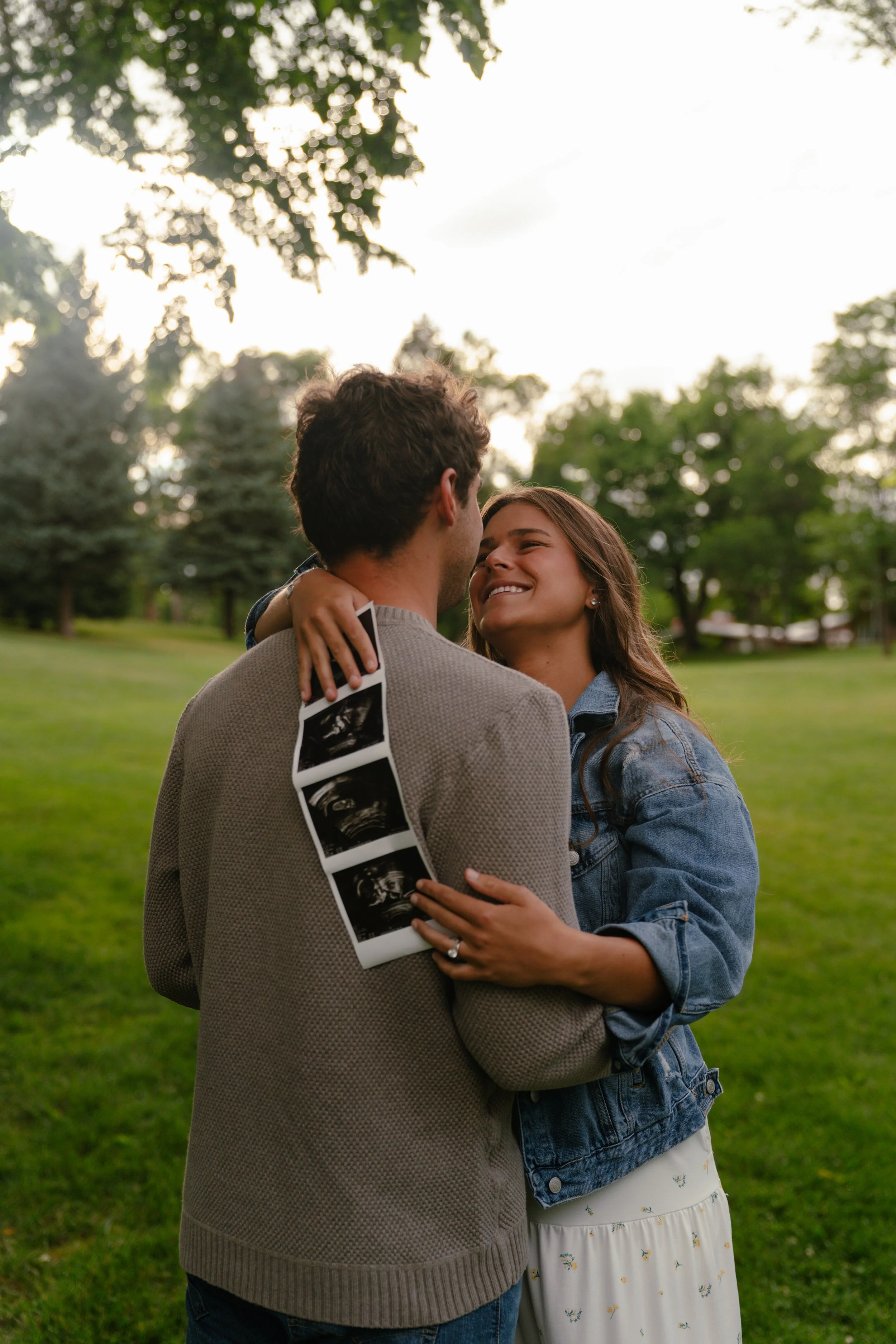 A couple embracing outdoors, woman smiling, holding ultrasound images, trees and bright sky in background.