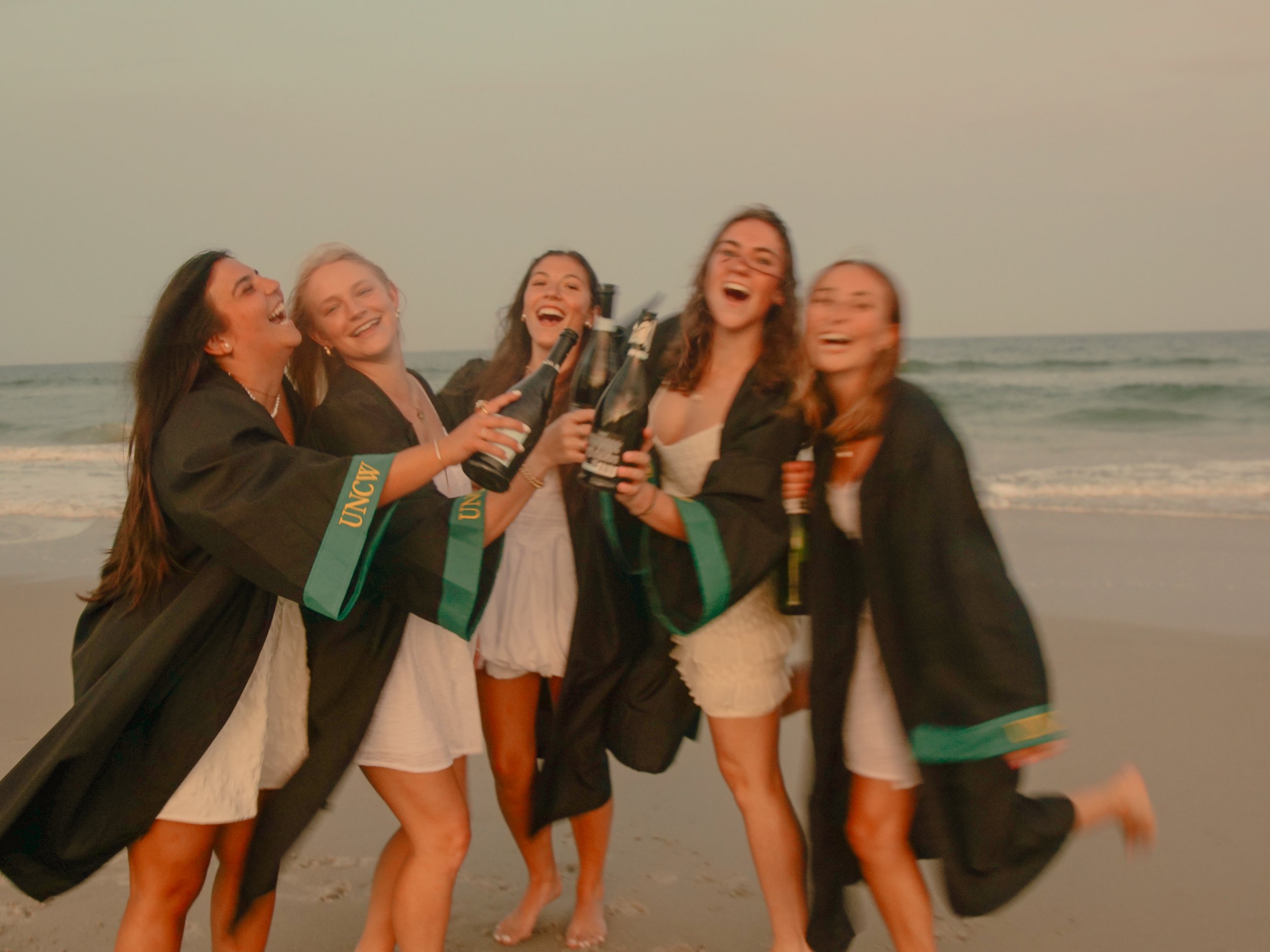 Group of women in graduation gowns celebrating on the beach, holding bottles of champagne, smiling and laughing.