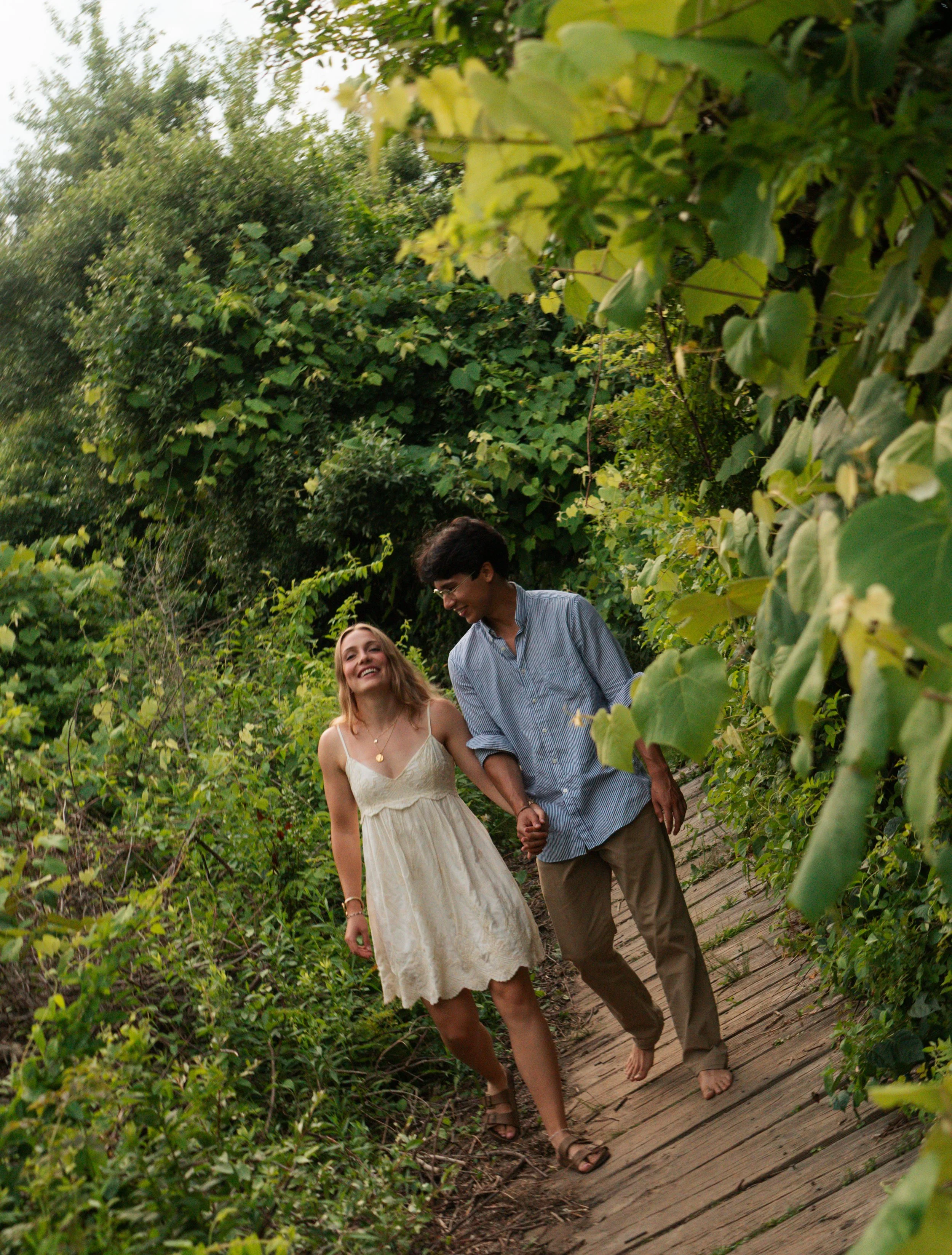 Two young adults holding hands and walking barefoot on a wooden path surrounded by lush green foliage, lawn, and trees in a natural setting.
