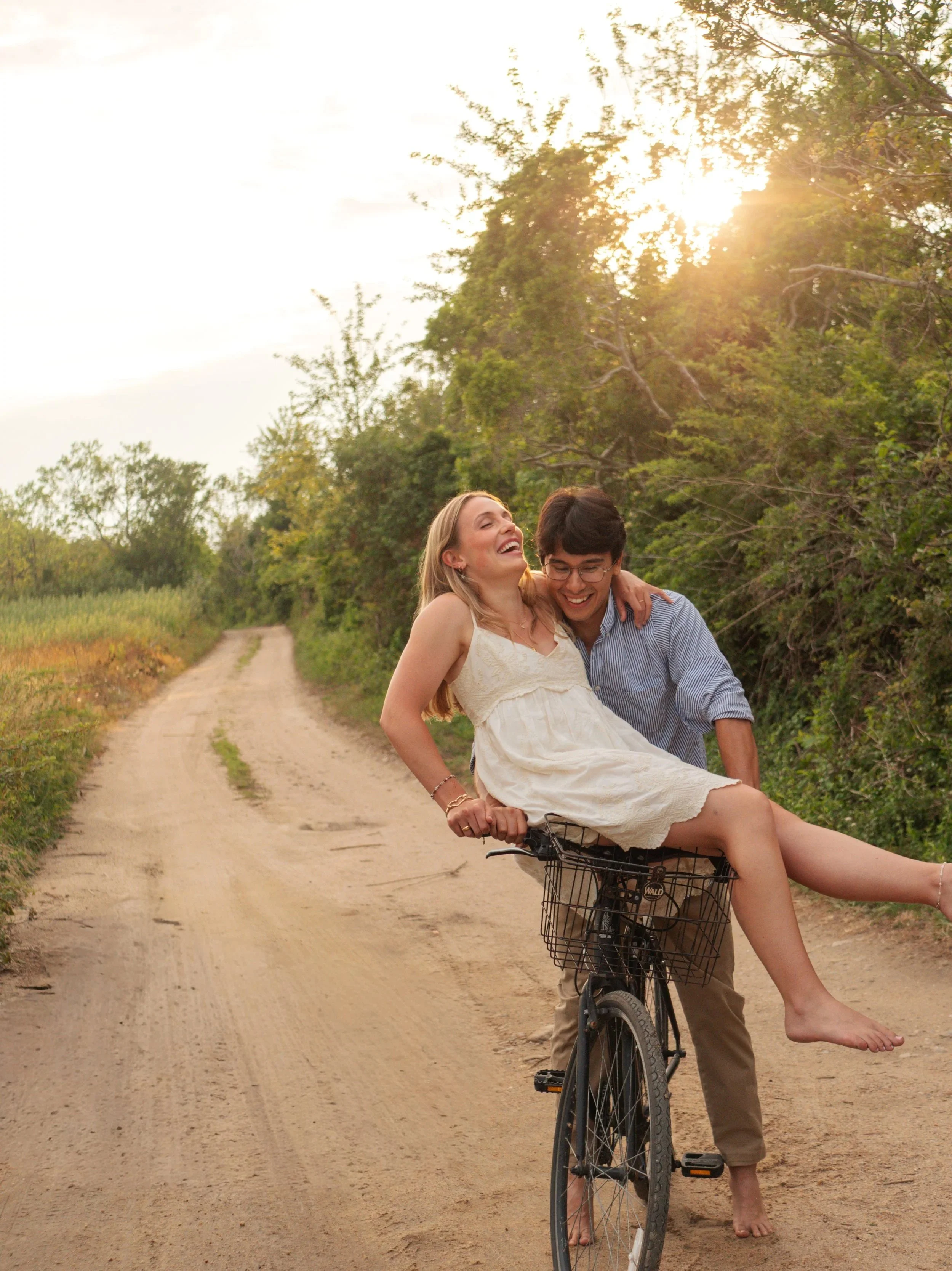 A man and woman enjoying a bike ride on a dirt path in nature, with the woman sitting on the front of the bicycle and the man holding her from behind, both smiling and laughing as the sun sets in the background.