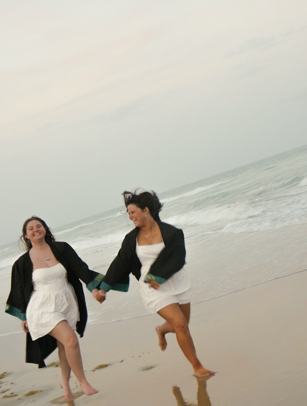 Two women in white dresses and black caps running barefoot on a beach holding hands, with ocean waves in the background.