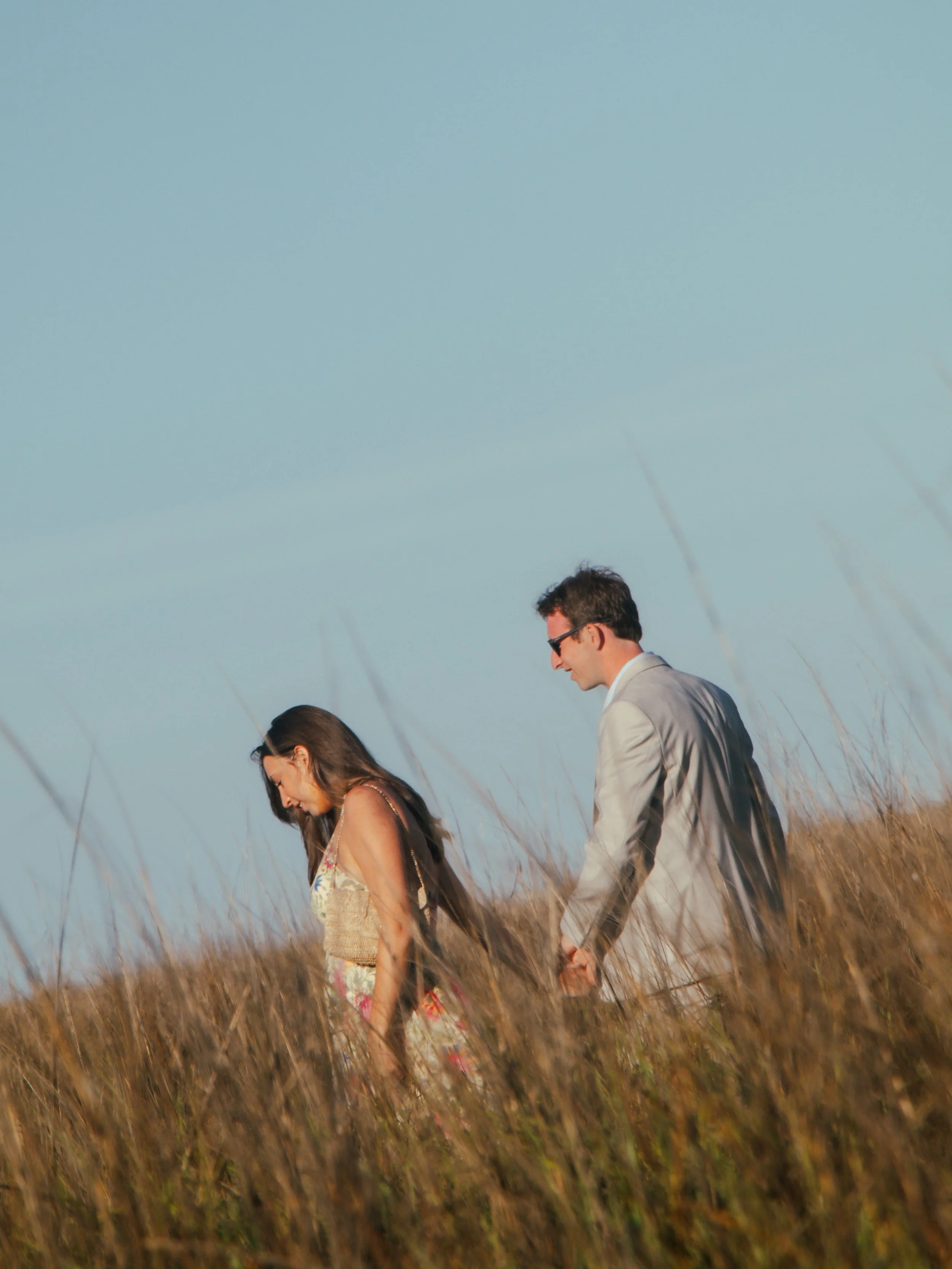 A man and a woman holding hands while walking through a field of tall, dry grass with a clear blue sky in the background.