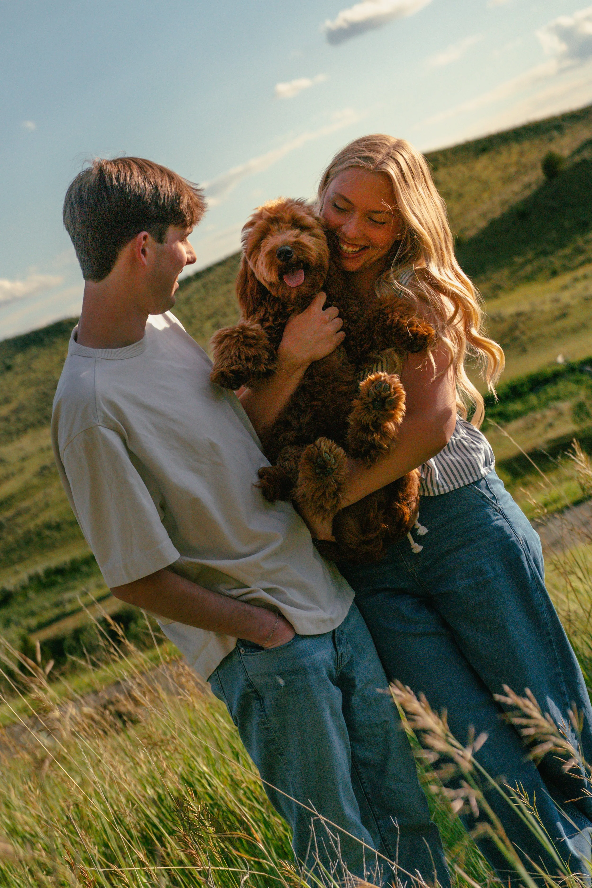 A young man and woman smiling while holding a happy puppy in a grassy field with rolling hills and blue sky in the background.